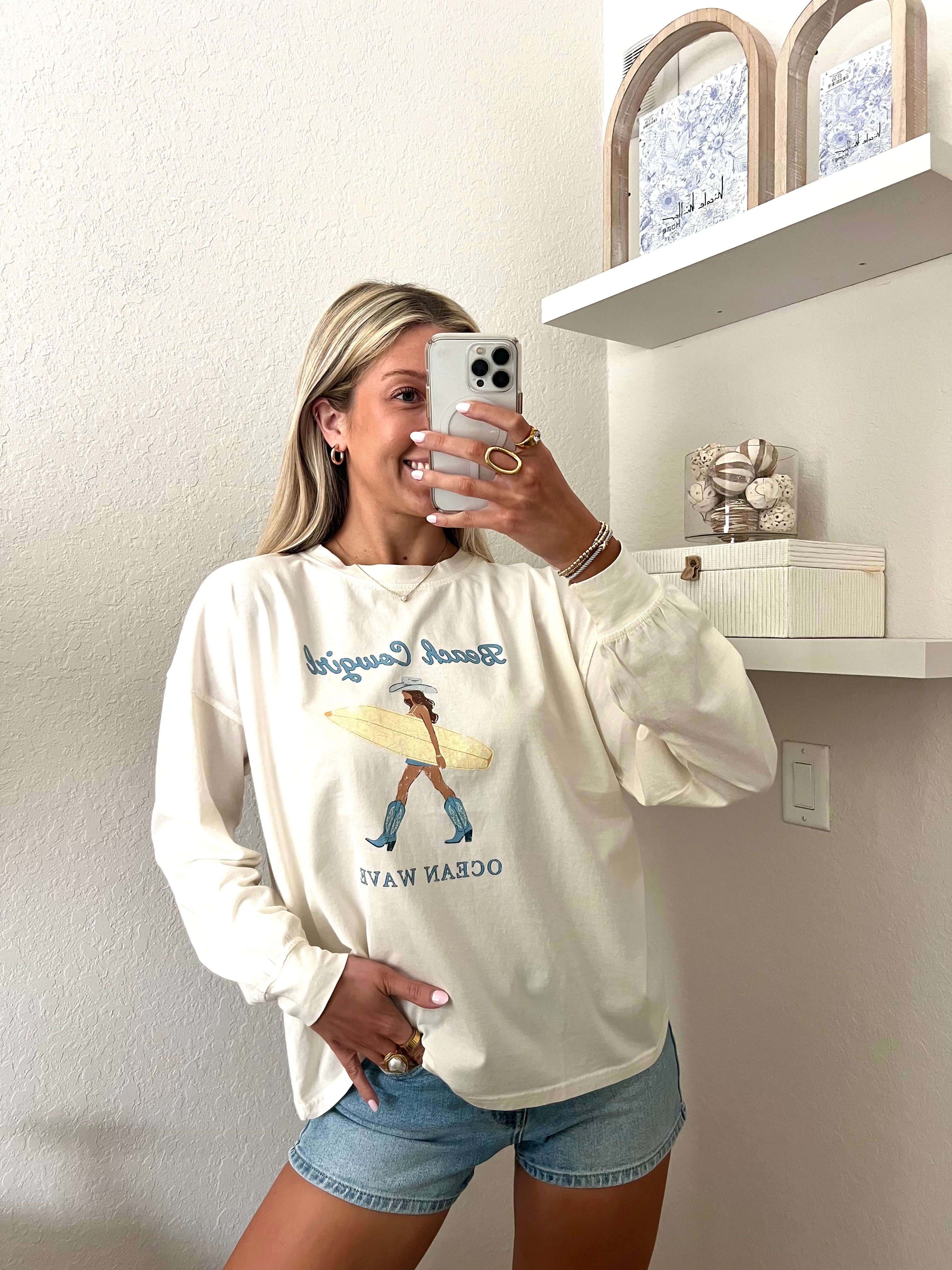 Woman wearing an ivory long sleeve tee with a beach cowgirl graphic design and denim shorts, taking a mirror selfie.