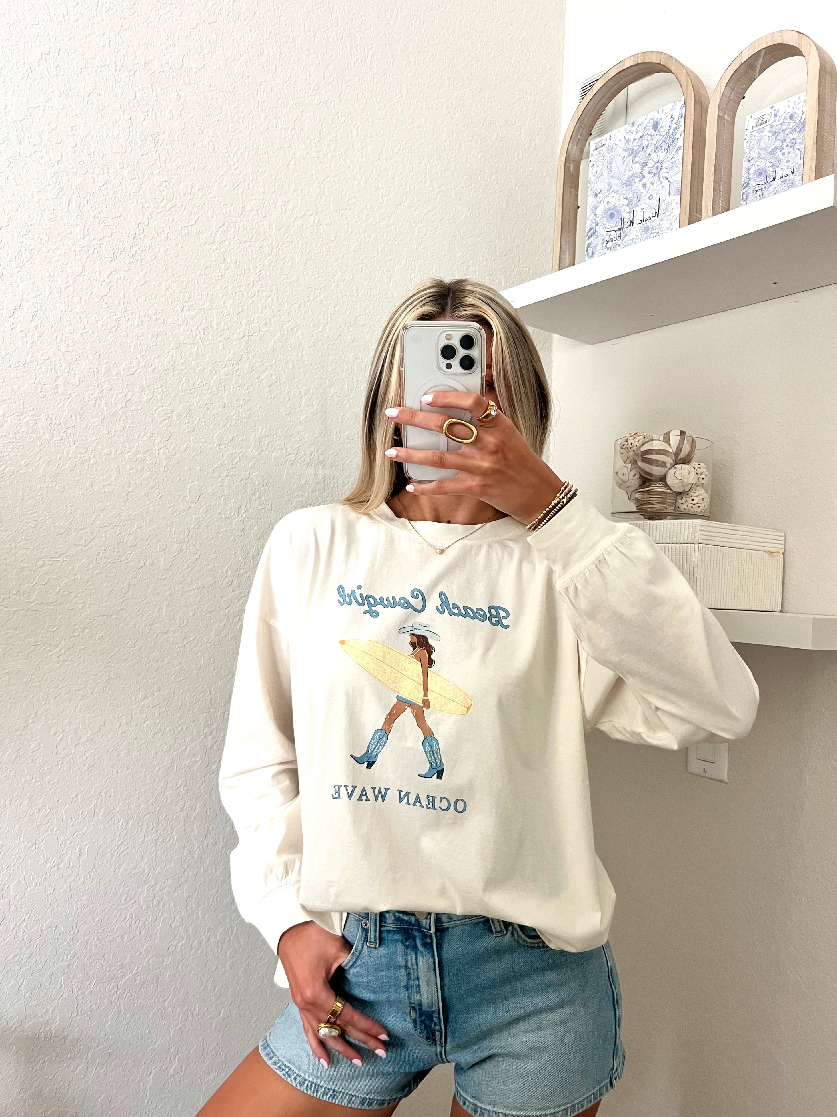 Woman wearing an ivory long sleeve tee with a beach cowgirl graphic design and denim shorts, taking a mirror selfie.