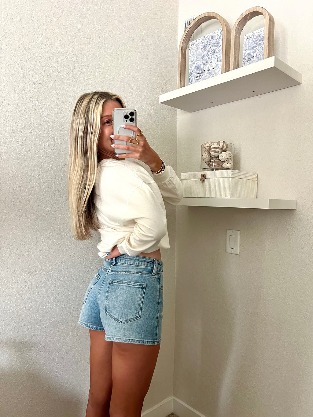 Woman taking a mirror selfie wearing light wash, high waisted denim shorts and a cream long sleeve shirt.