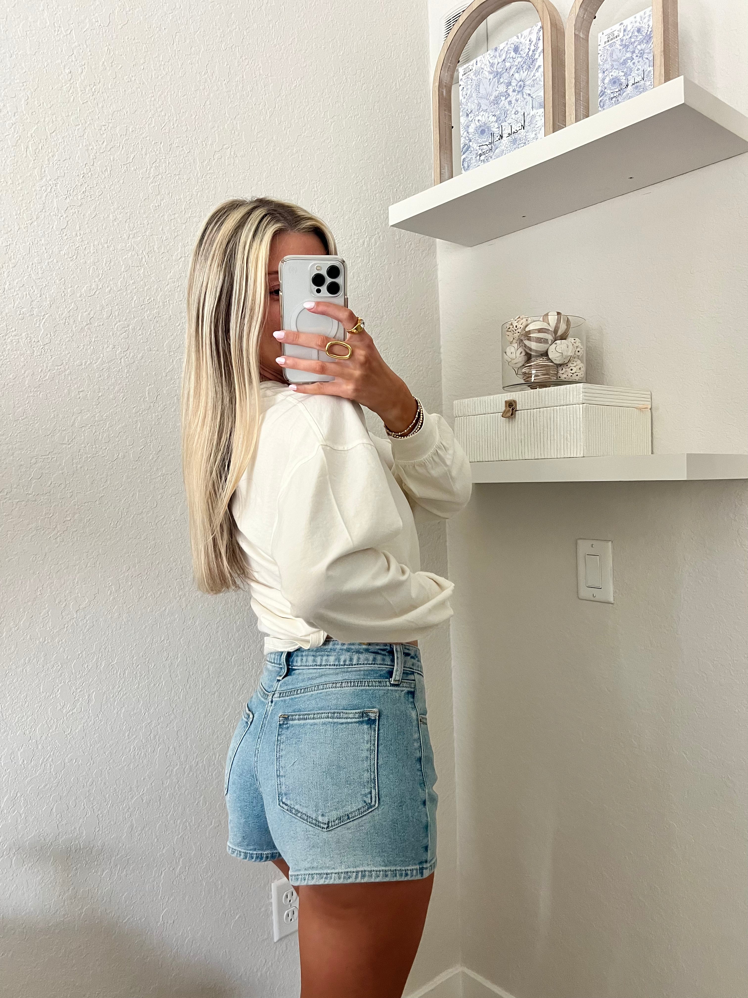 Woman taking a mirror selfie wearing light wash, high waisted denim shorts and a cream long sleeve shirt.
