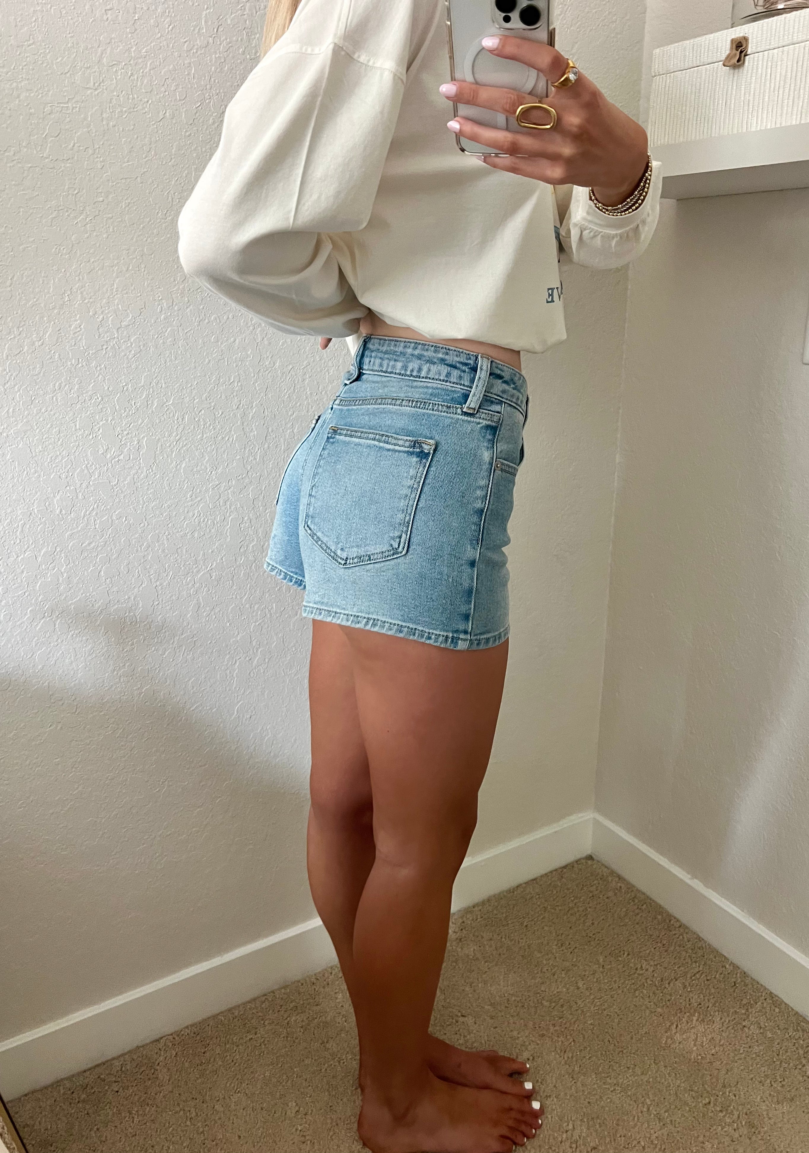 Woman taking a mirror selfie wearing light wash, high waisted denim shorts and a cream long sleeve shirt.