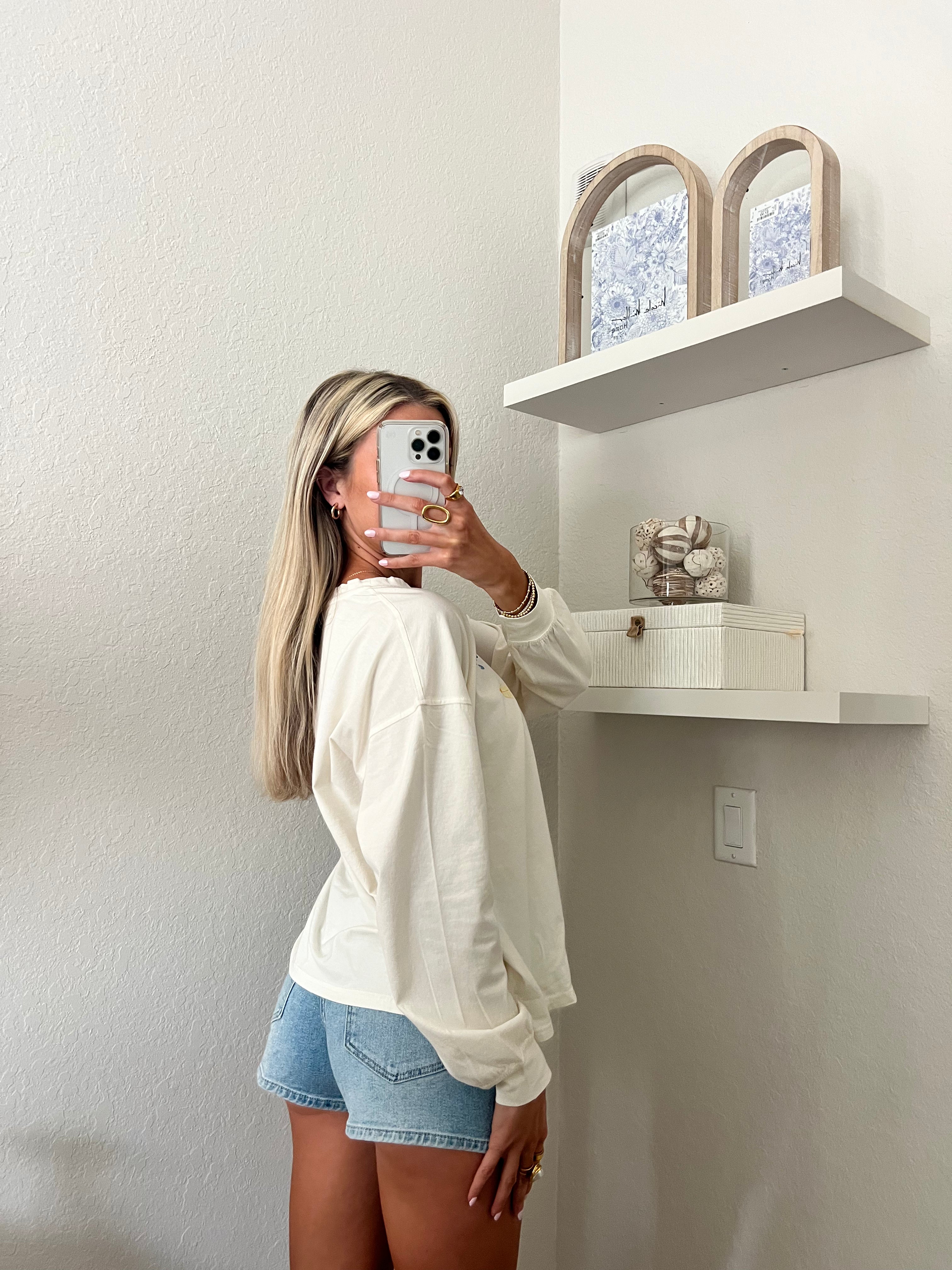 Woman wearing an ivory long sleeve tee with a beach cowgirl graphic design and denim shorts, taking a mirror selfie.