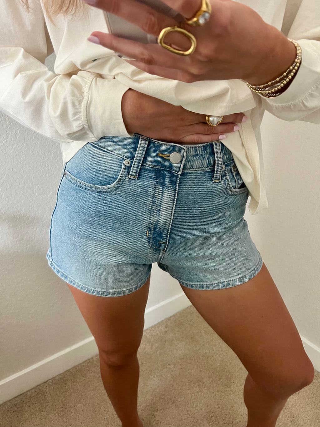 Woman taking a mirror selfie wearing light wash, high waisted denim shorts and a cream long sleeve shirt.
