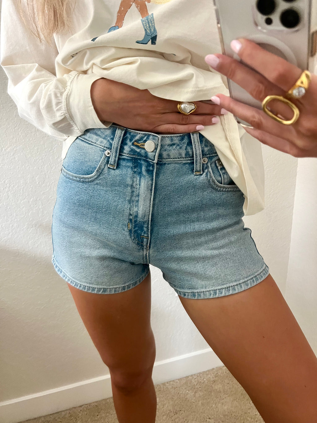 Woman taking a mirror selfie wearing light wash, high waisted denim shorts.