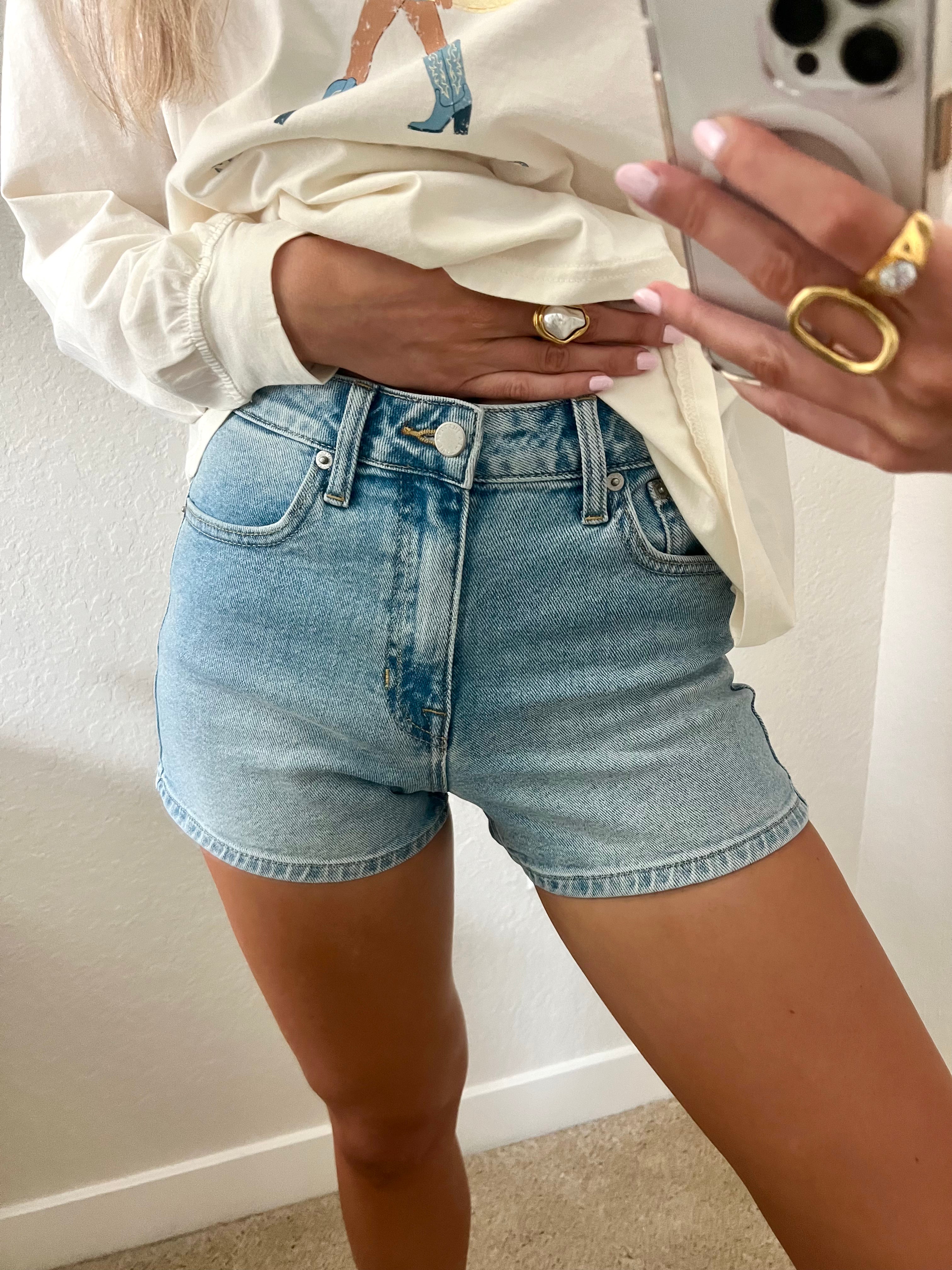 Woman taking a mirror selfie wearing light wash, high waisted denim shorts.