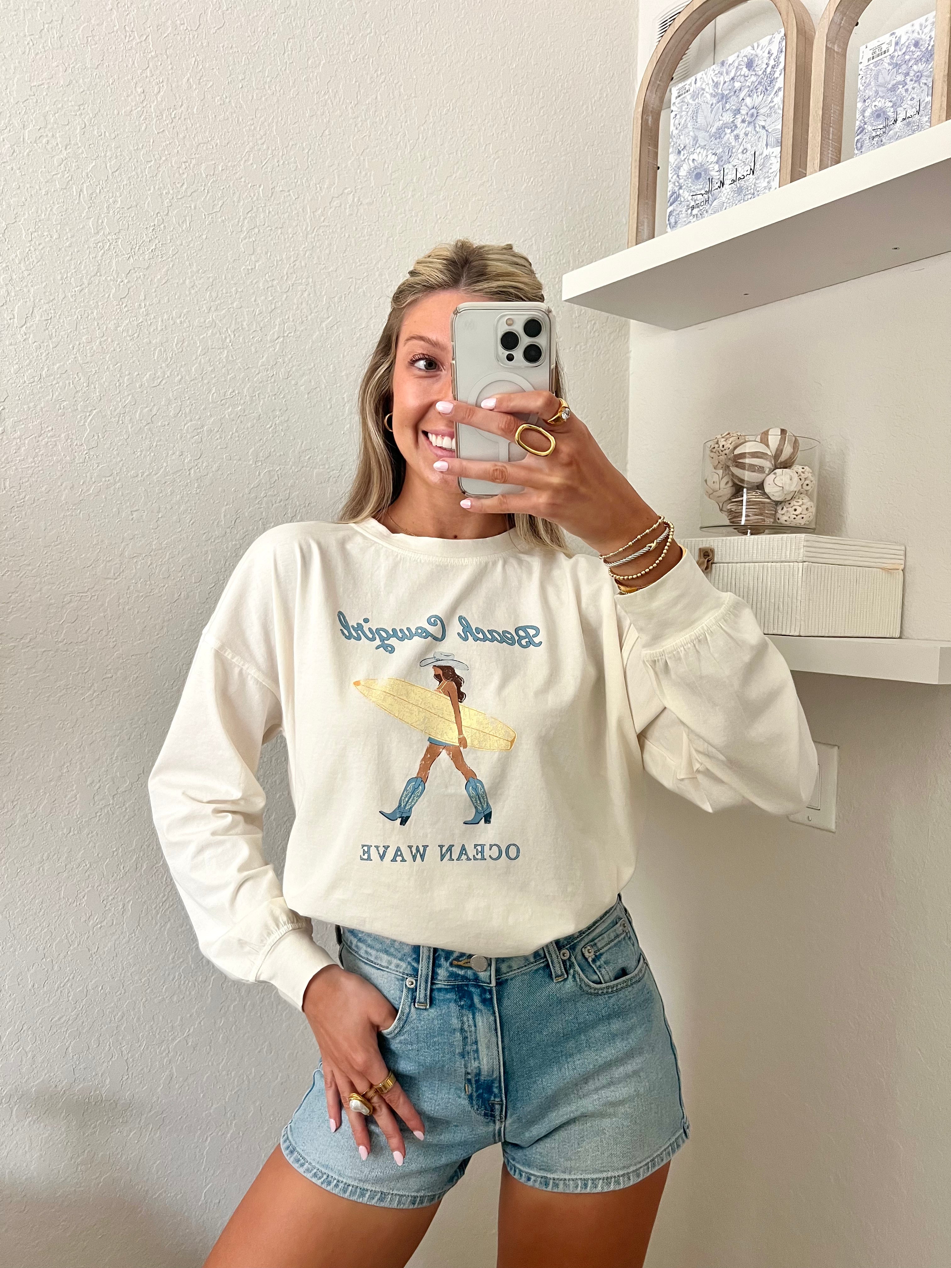 Woman wearing an ivory long sleeve tee with a beach cowgirl graphic design and denim shorts, taking a mirror selfie.