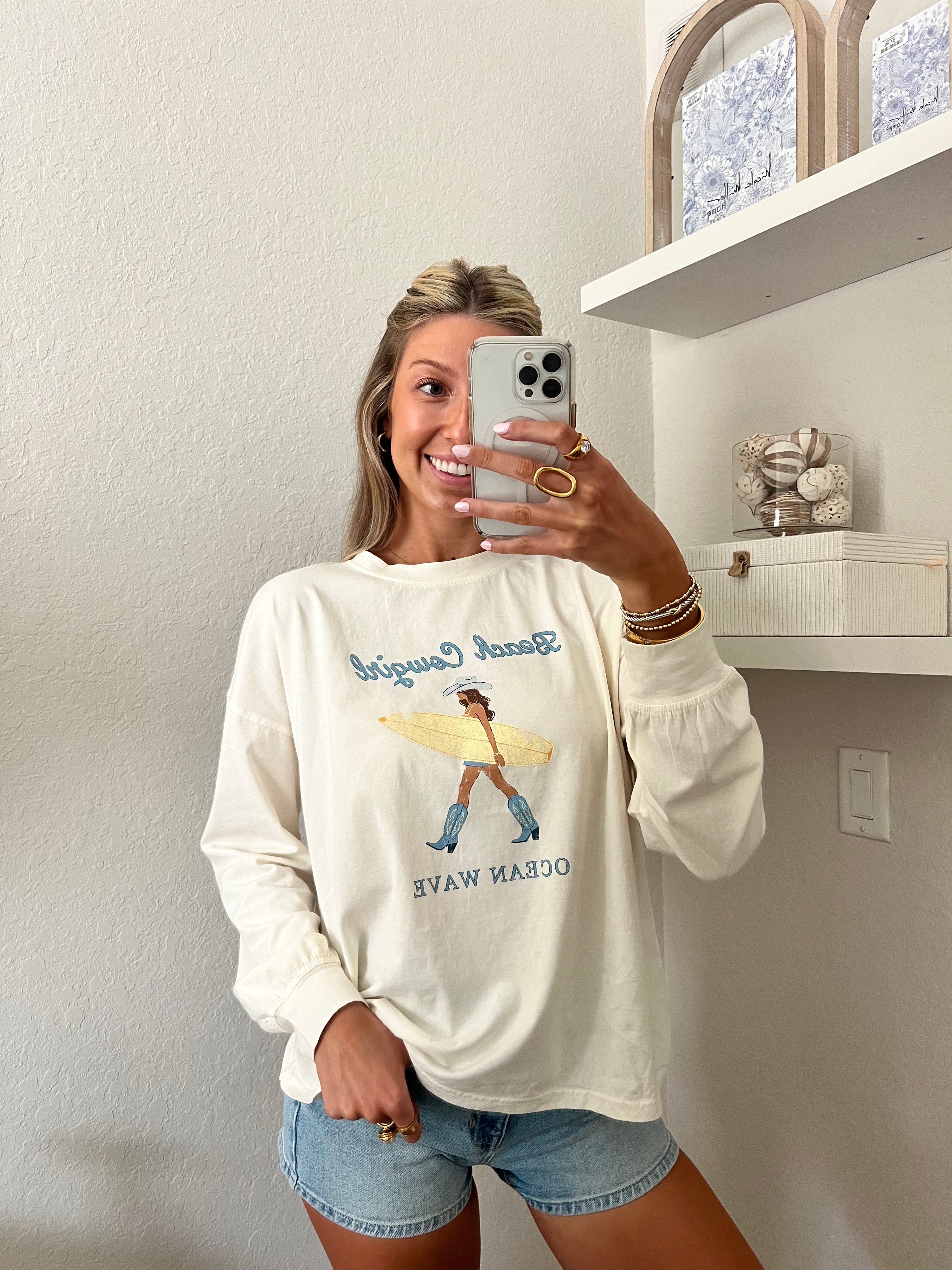 Woman wearing an ivory long sleeve tee with a beach cowgirl graphic design and denim shorts, taking a mirror selfie.