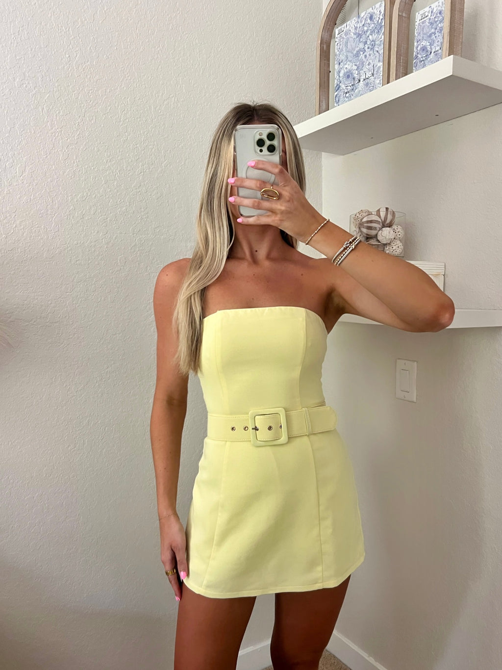 Woman taking a mirror selfie wearing a yellow strapless belted dress in a minimalist room.