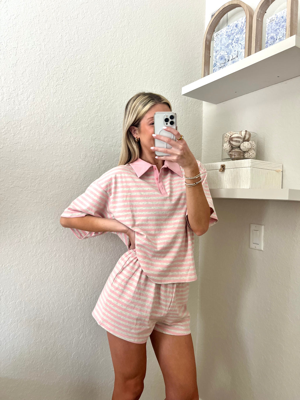 Woman taking a mirror selfie wearing a pink and grey striped short sleeve collared shirt and shorts set in a room with a shelf and decorative items.