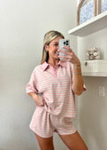 Woman taking a mirror selfie wearing a pink and grey striped short sleeve collared shirt and shorts set in a room with a shelf and decorative items.
