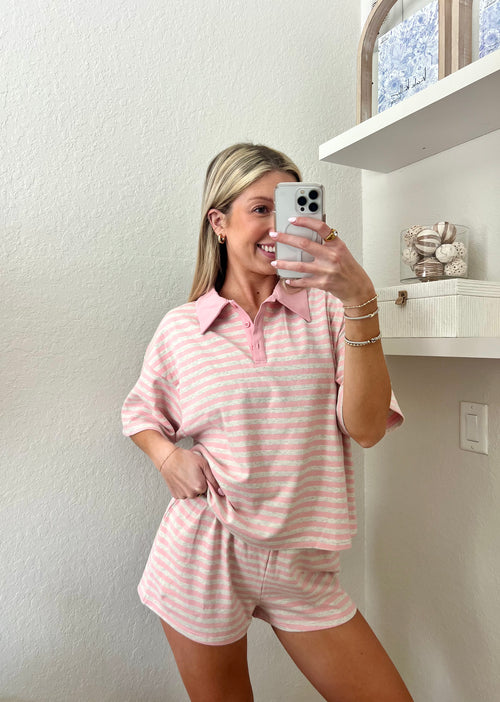 Woman taking a mirror selfie wearing a pink and grey striped short sleeve collared shirt and shorts set in a room with a shelf and decorative items.