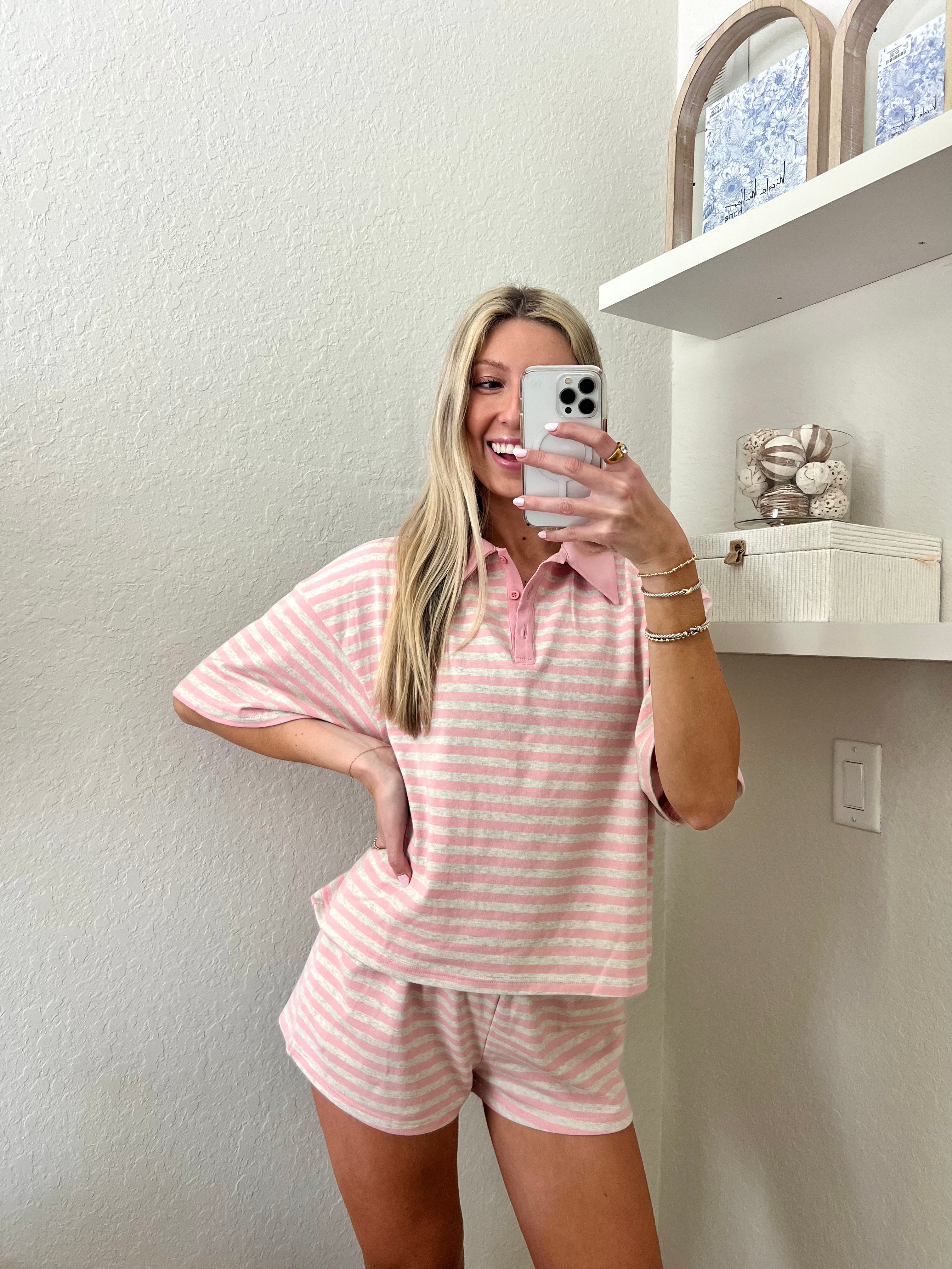Woman taking a mirror selfie wearing a pink and grey striped short sleeve collared shirt and shorts set in a room with a shelf and decorative items.