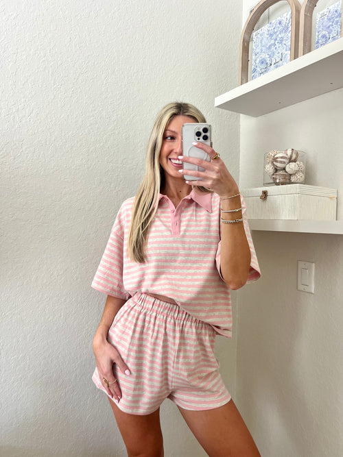 Woman taking a mirror selfie wearing a pink and grey striped short sleeve collared shirt and shorts set in a room with a shelf and decorative items.