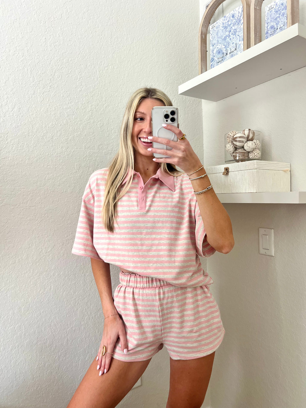 Woman taking a mirror selfie wearing a pink and grey striped short sleeve collared shirt and shorts set in a room with a shelf and decorative items.