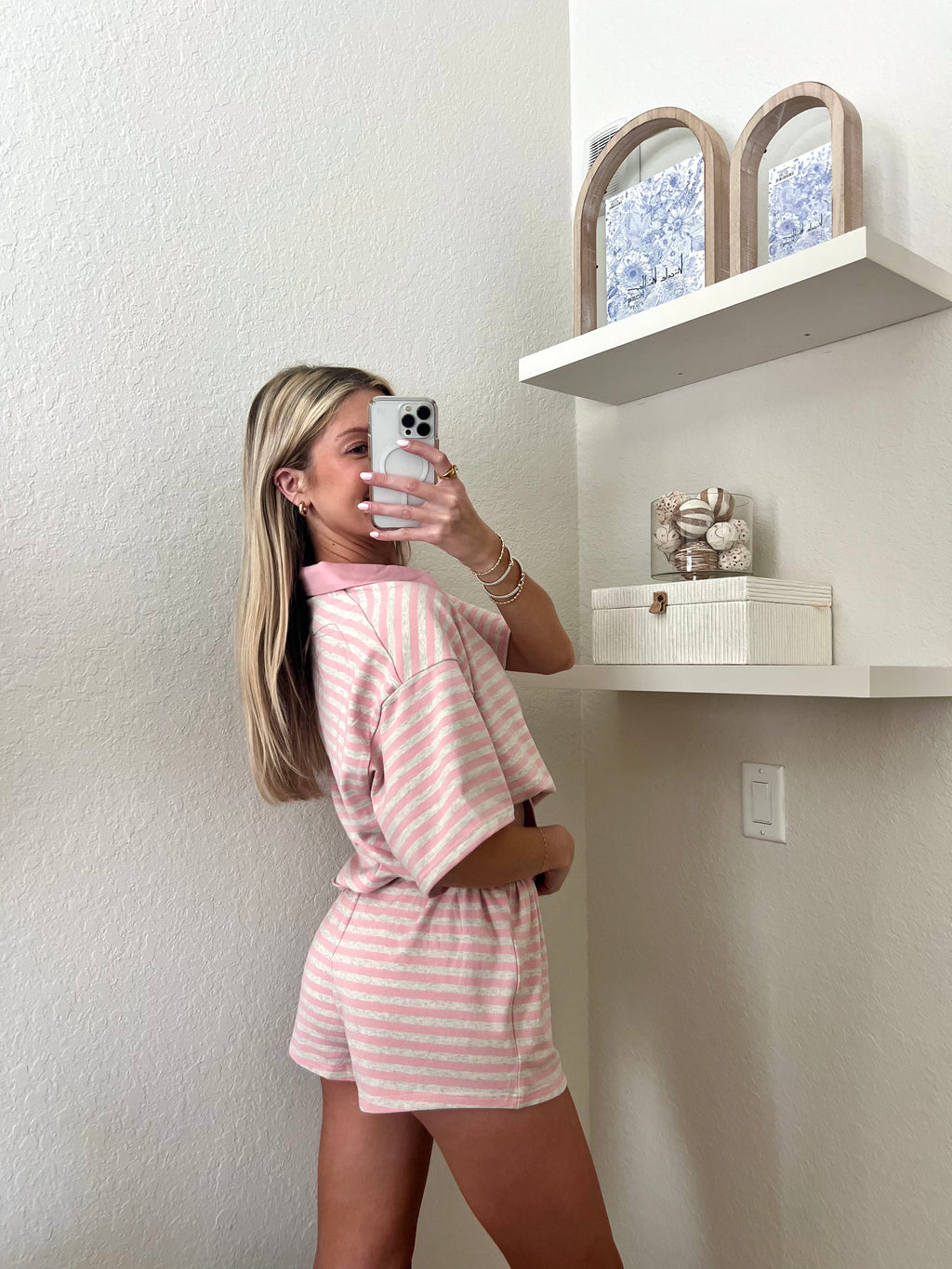 Woman taking a mirror selfie wearing a pink and grey striped short sleeve collared shirt and shorts set in a room with a shelf and decorative items.