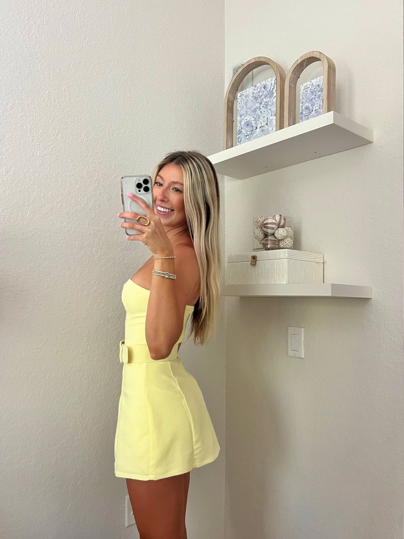 Woman taking a mirror selfie wearing a yellow strapless belted dress in a minimalist room.