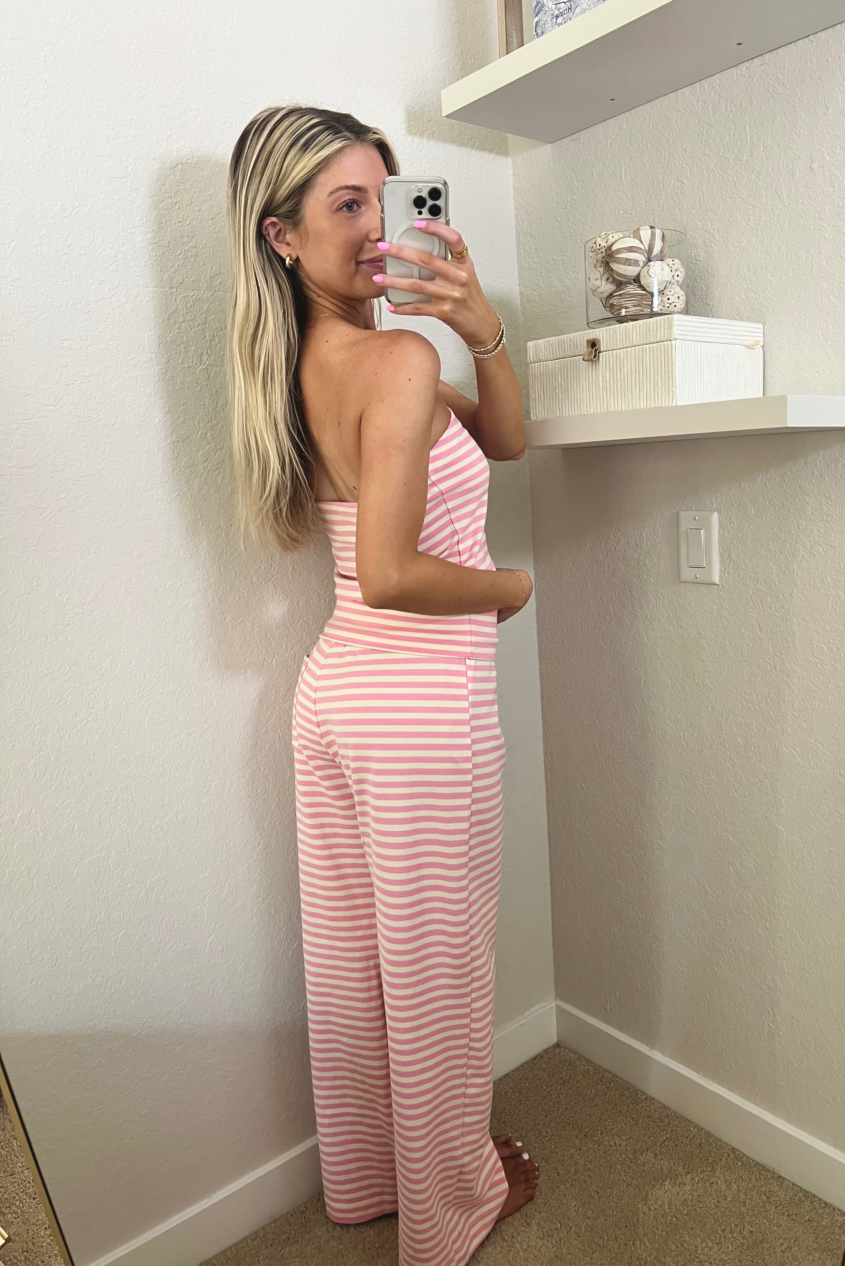 Woman taking a mirror selfie wearing a pink and ivory striped tube top and pants set in a room.
