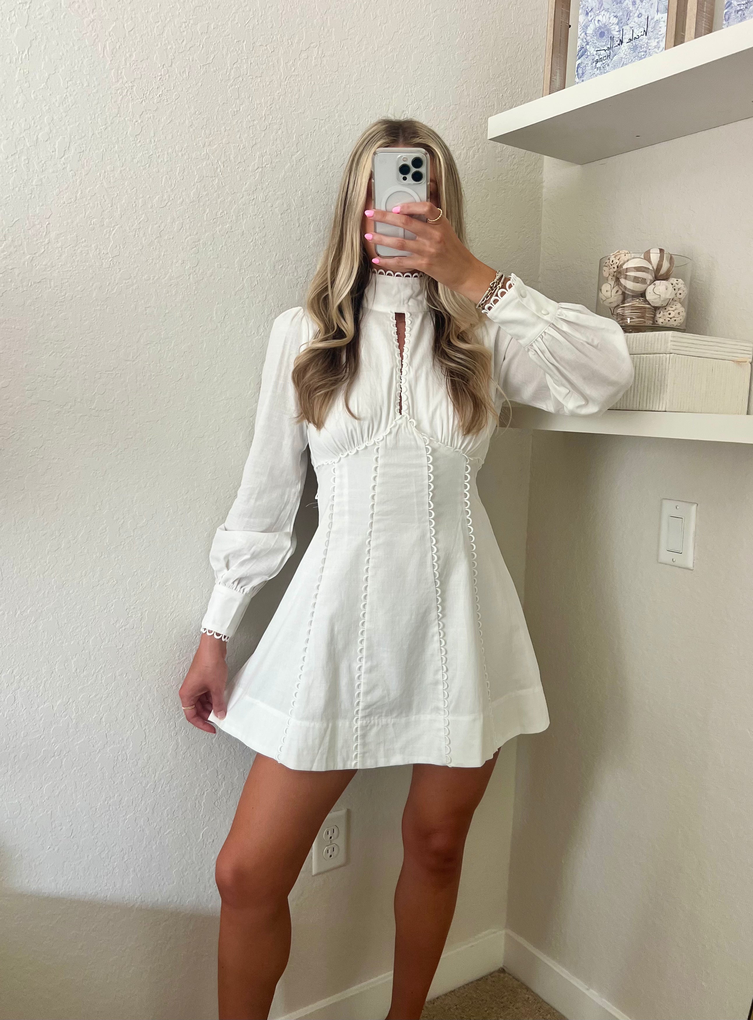 Woman taking a mirror selfie wearing a white long sleeve mini dress in a room with a shelf and decorative items.