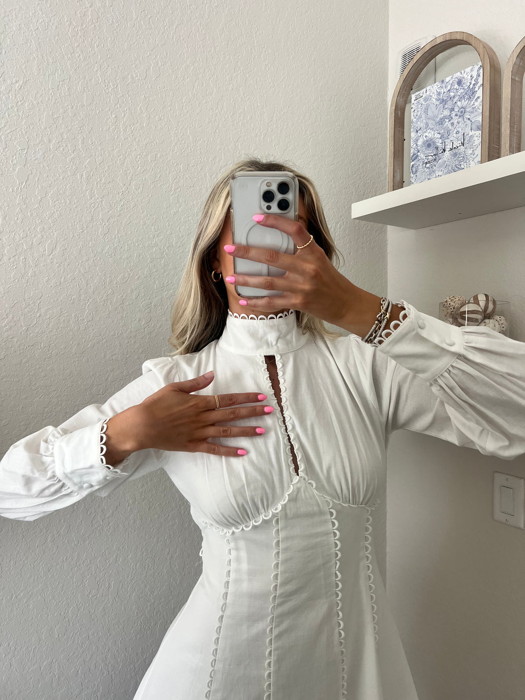 Woman taking a mirror selfie wearing a white long sleeve mini dress in a room with a shelf and decorative items.