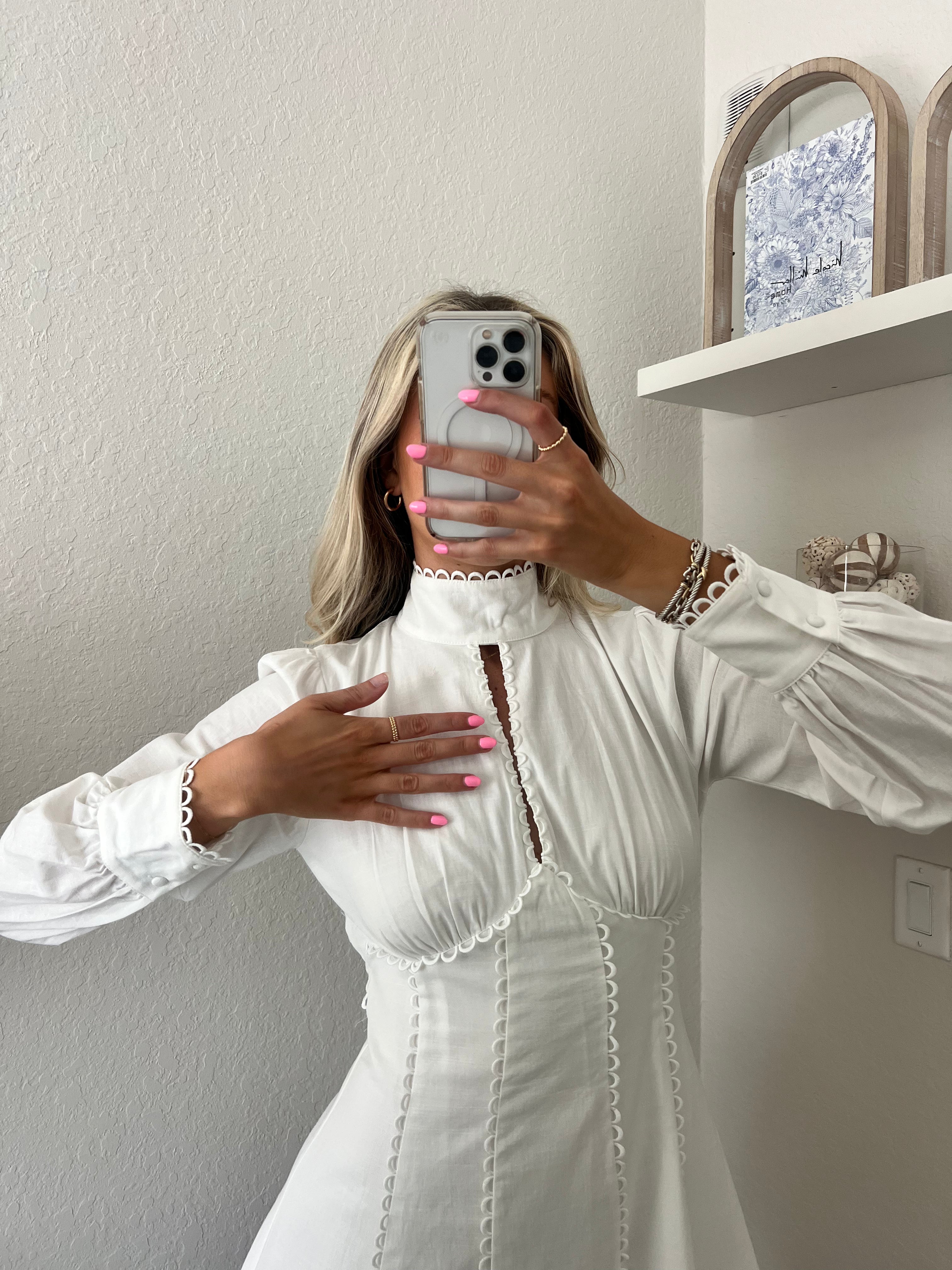 Woman taking a mirror selfie wearing a white long sleeve mini dress in a room with a shelf and decorative items.