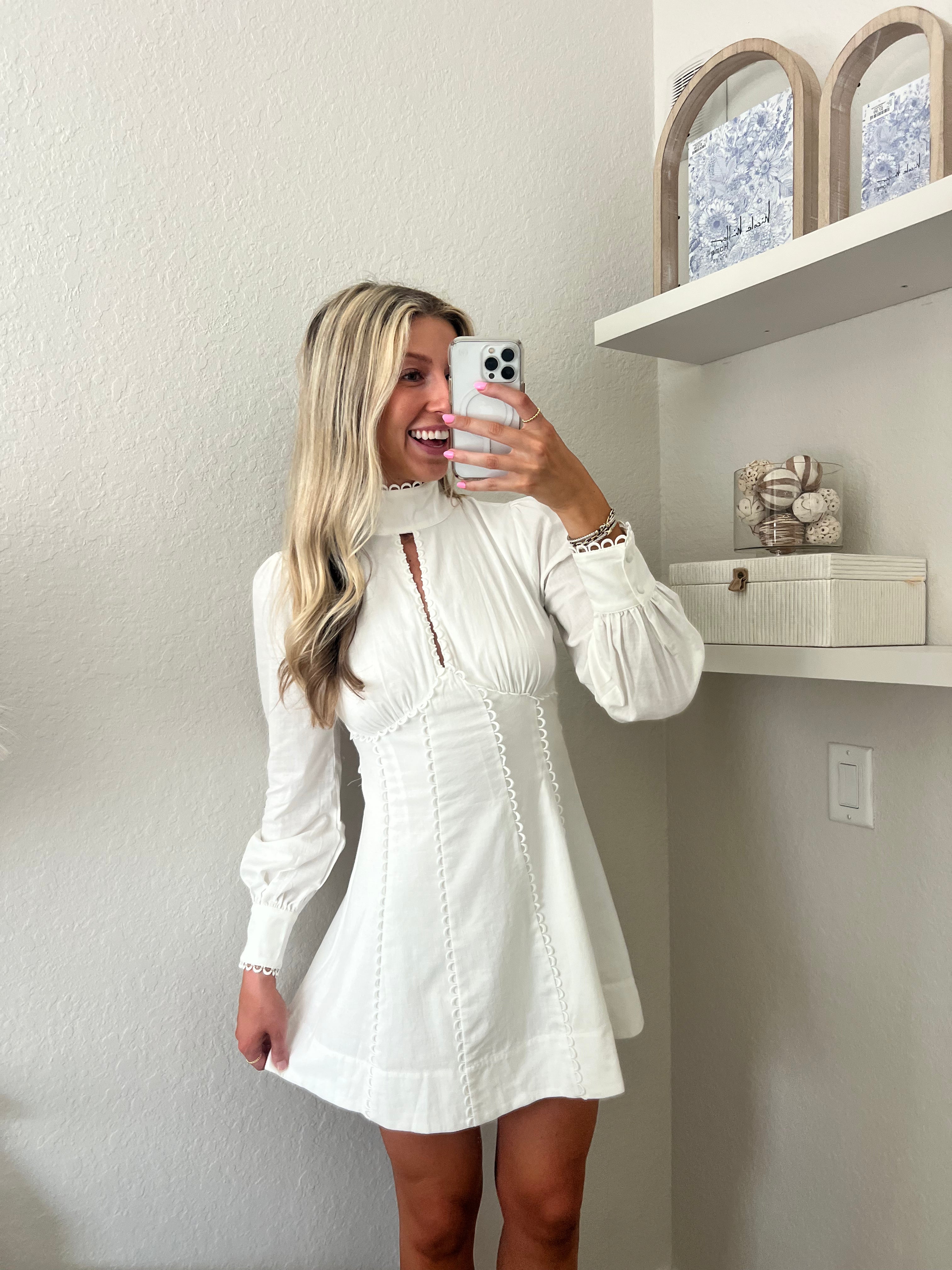 Woman taking a mirror selfie wearing a white long sleeve mini dress in a room with a shelf and decorative items.