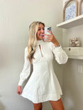 Woman taking a mirror selfie wearing a white long sleeve mini dress in a room with a shelf and decorative items.