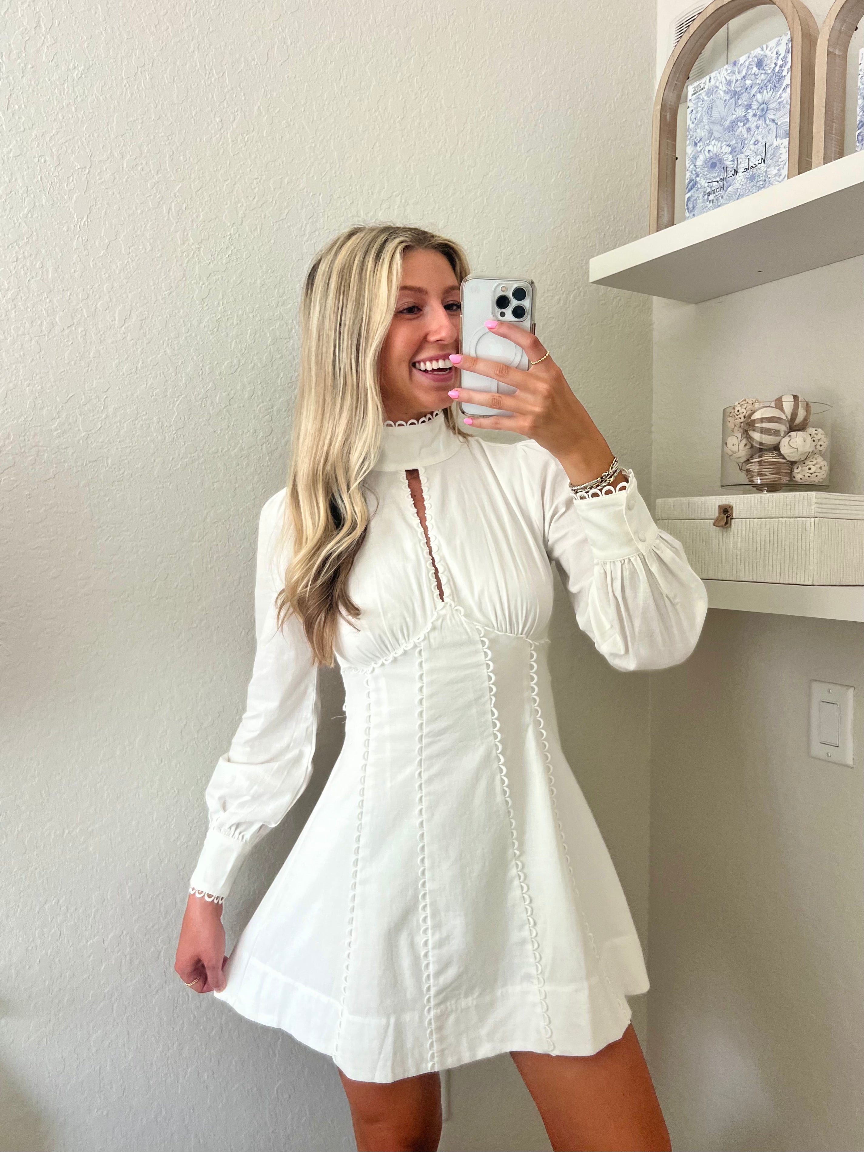 Woman taking a mirror selfie wearing a white long sleeve mini dress in a room with a shelf and decorative items.