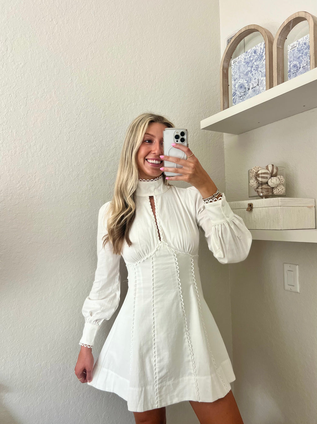 Woman taking a mirror selfie wearing a white long sleeve mini dress in a room with a shelf and decorative items.