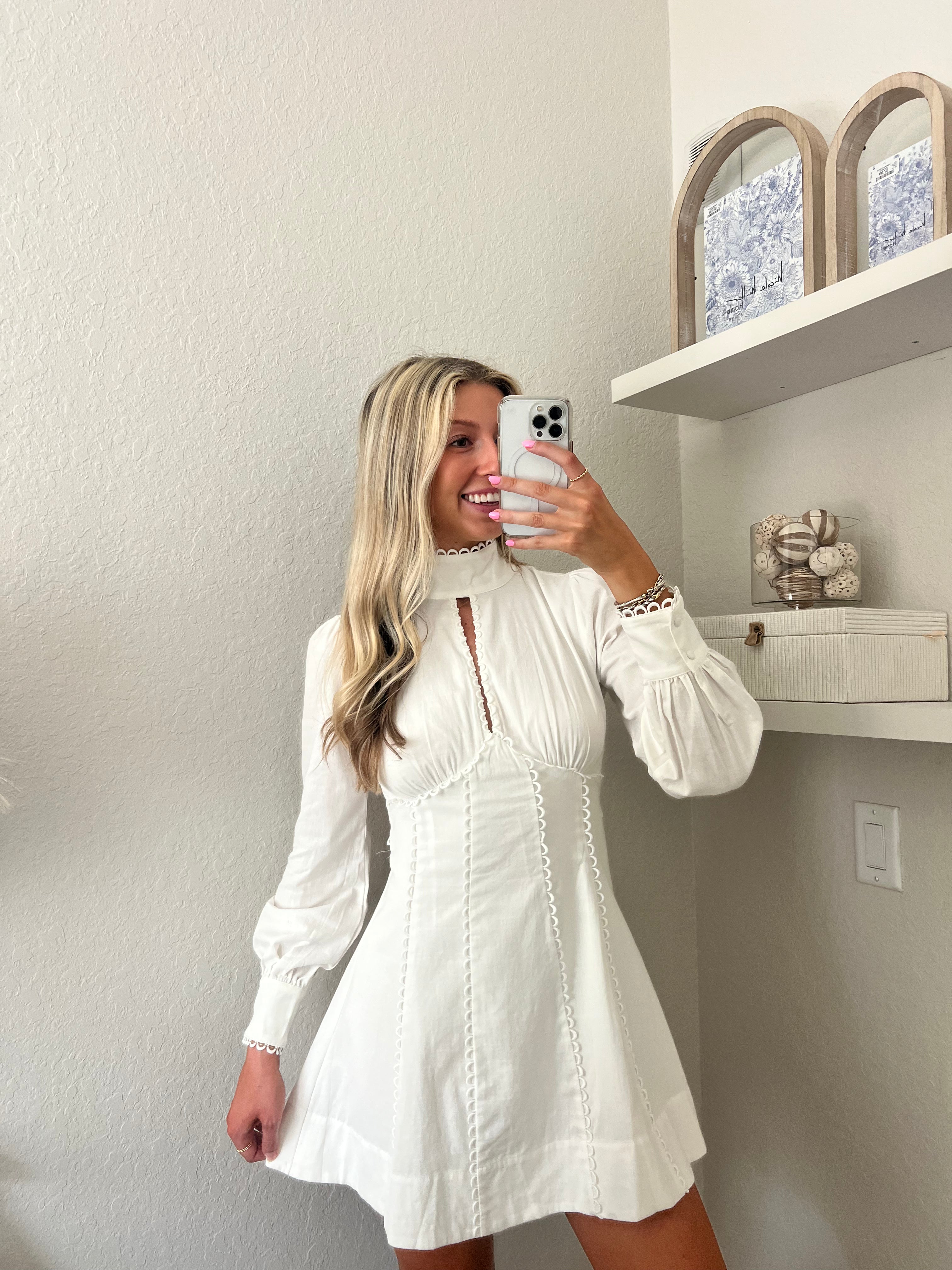 Woman taking a mirror selfie wearing a white long sleeve mini dress in a room with a shelf and decorative items.