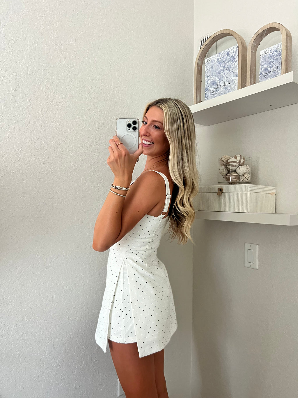 Woman taking a mirror selfie wearing a white dress with small black polka dots in a room with a shelf and decorative items.