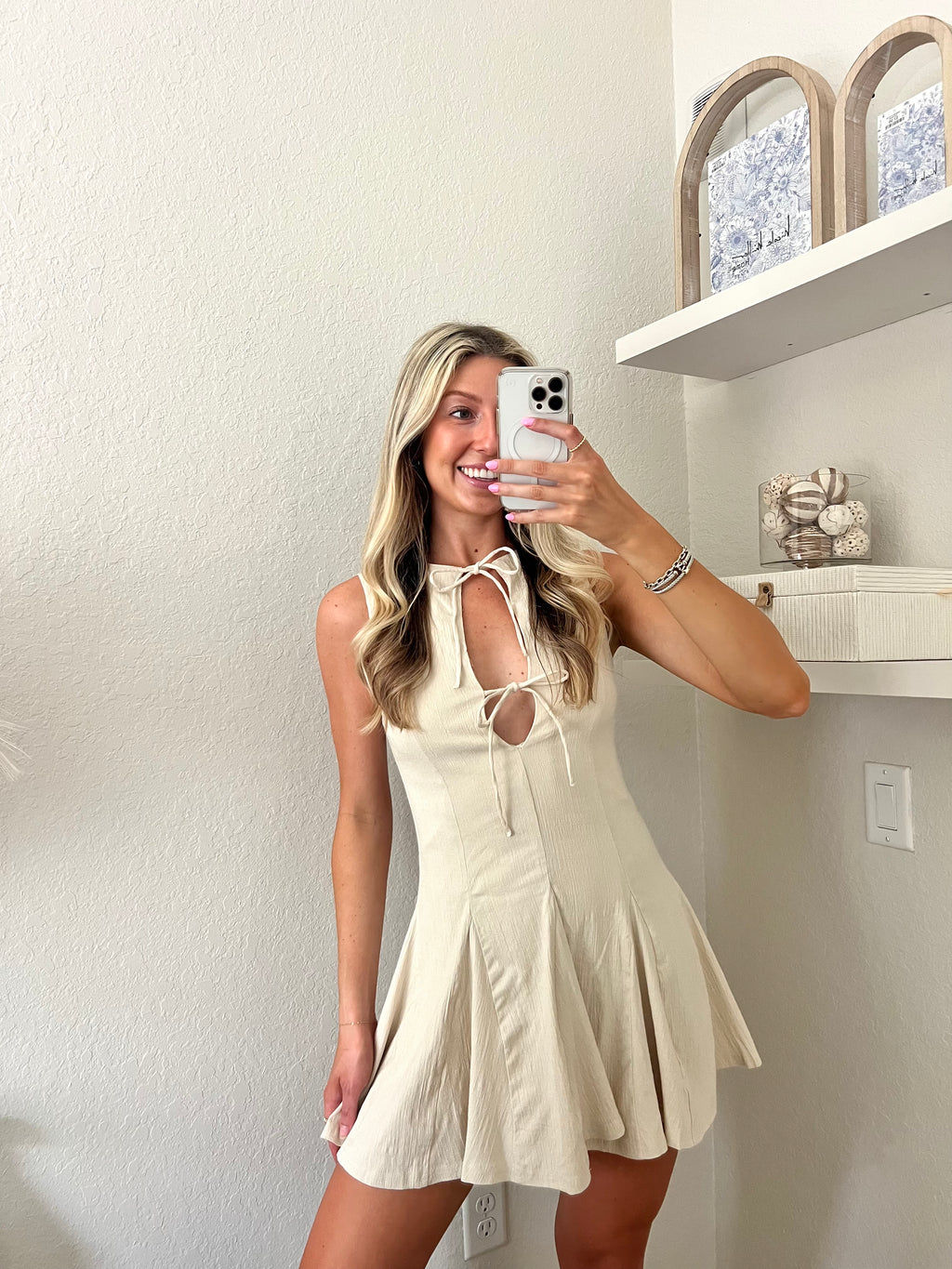 Woman taking a mirror selfie wearing a cream neutral dress with bows on the chest in a room with white walls and a shelf.