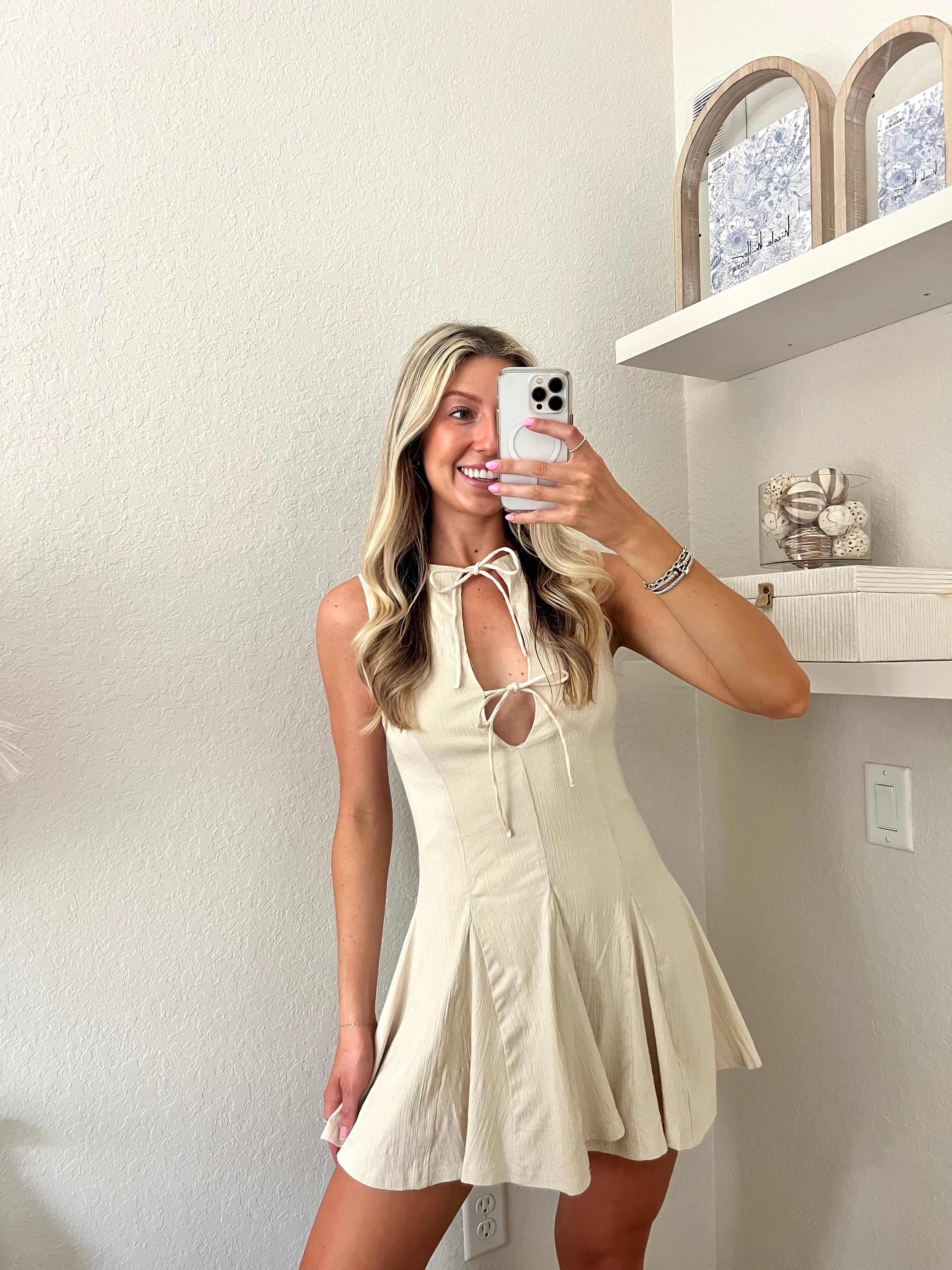 Woman taking a mirror selfie wearing a cream neutral dress with bows on the chest in a room with white walls and a shelf.