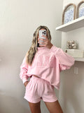 Woman wearing a pink sweatshirt with the word "Salty" on it and shorts set in a room with a shelf in the background.
