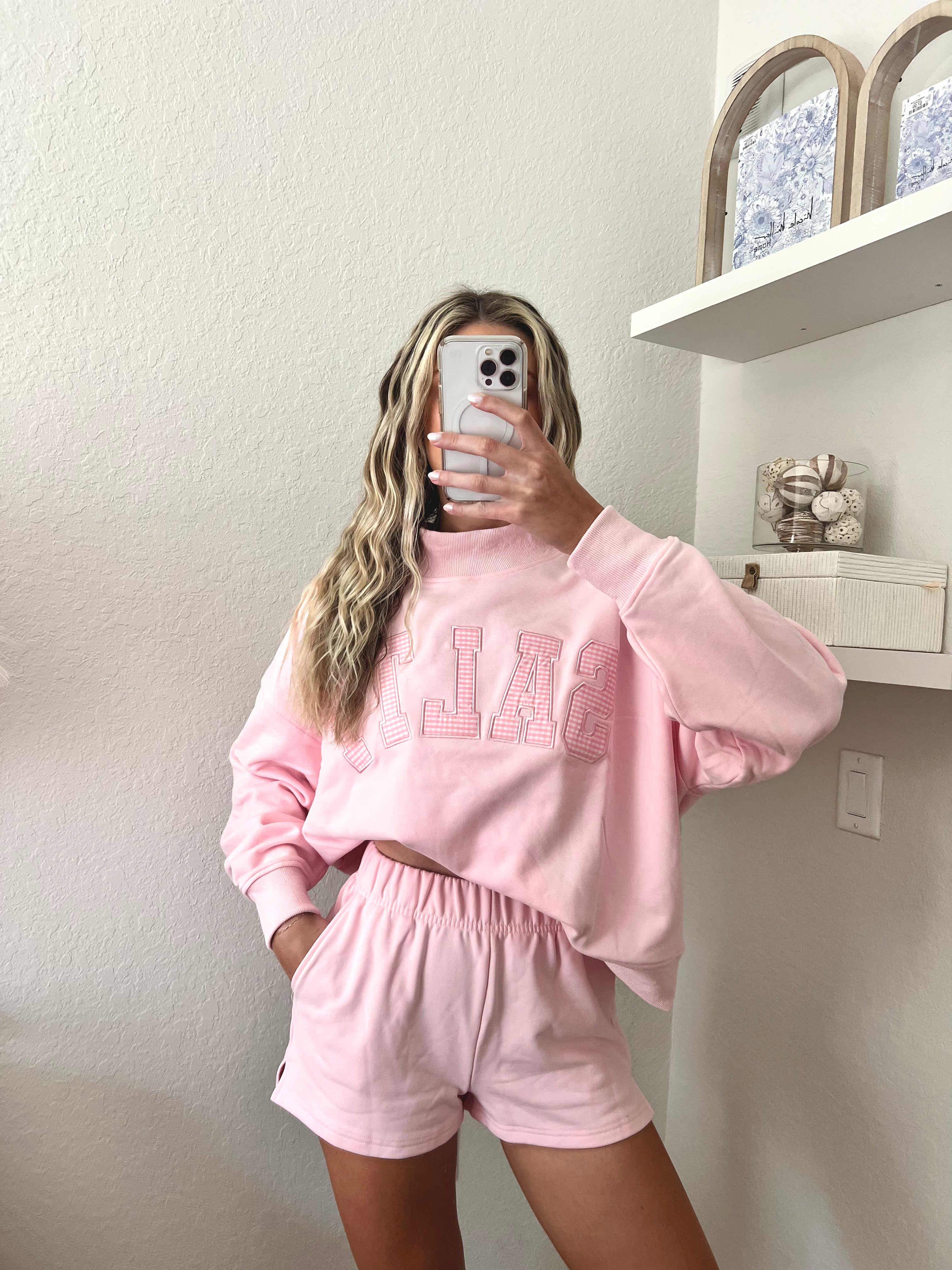 Woman wearing a pink sweatshirt with the word "Salty" on it and shorts set in a room with a shelf in the background.
