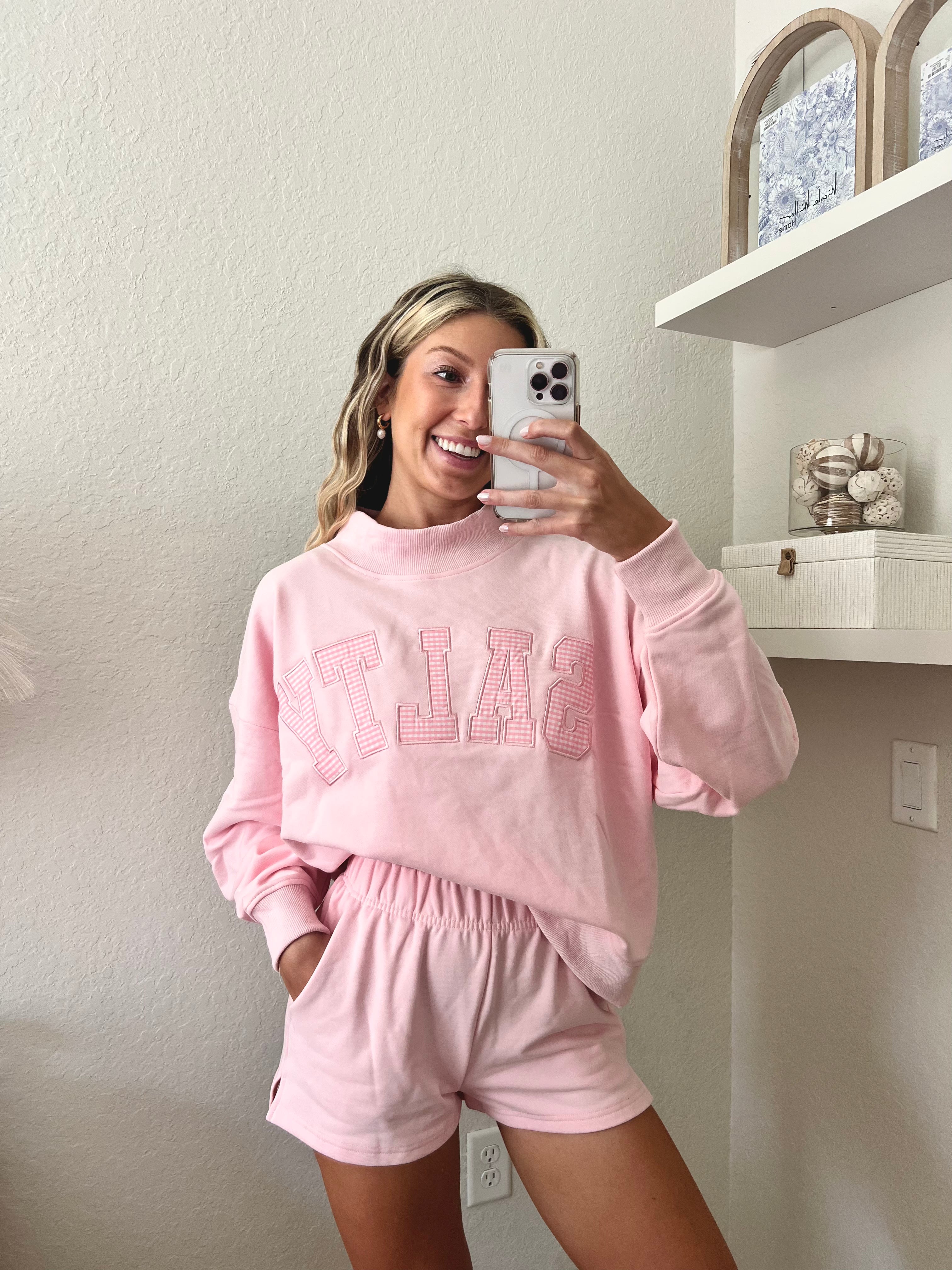 Woman wearing a pink sweatshirt with the word "Salty" on it and shorts set in a room with a shelf in the background.