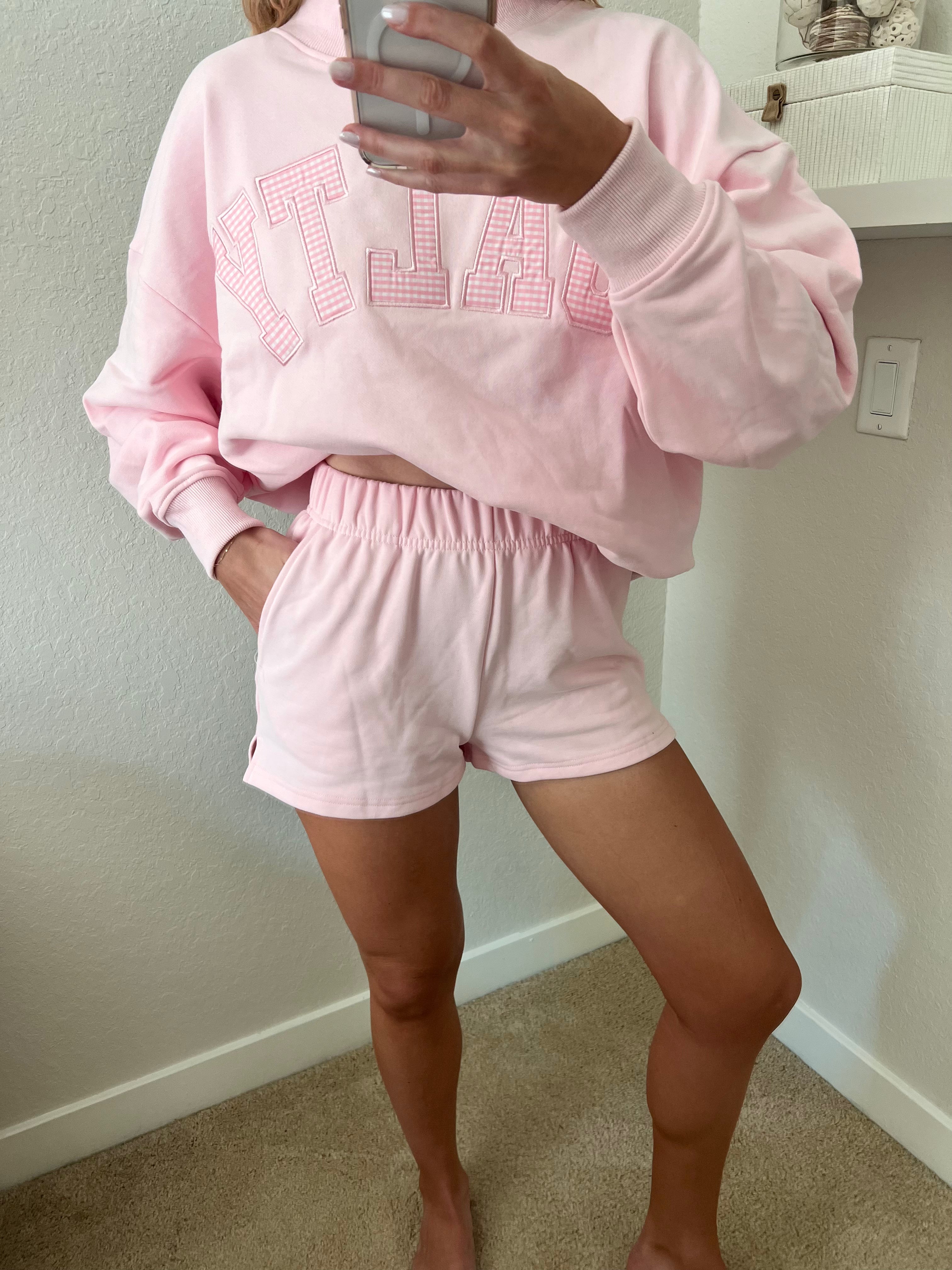 Woman wearing a pink sweatshirt with the word "Salty" on it and shorts set in a room with a shelf in the background.