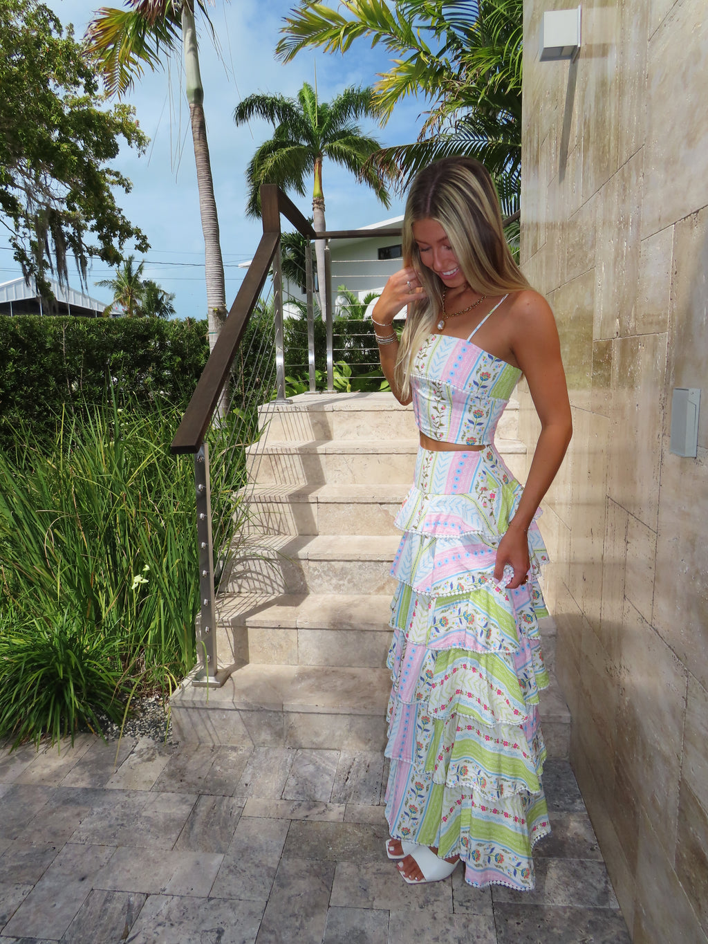 Woman in a patterned maxi skirt set standing against a stone wall with trees and sky in the background