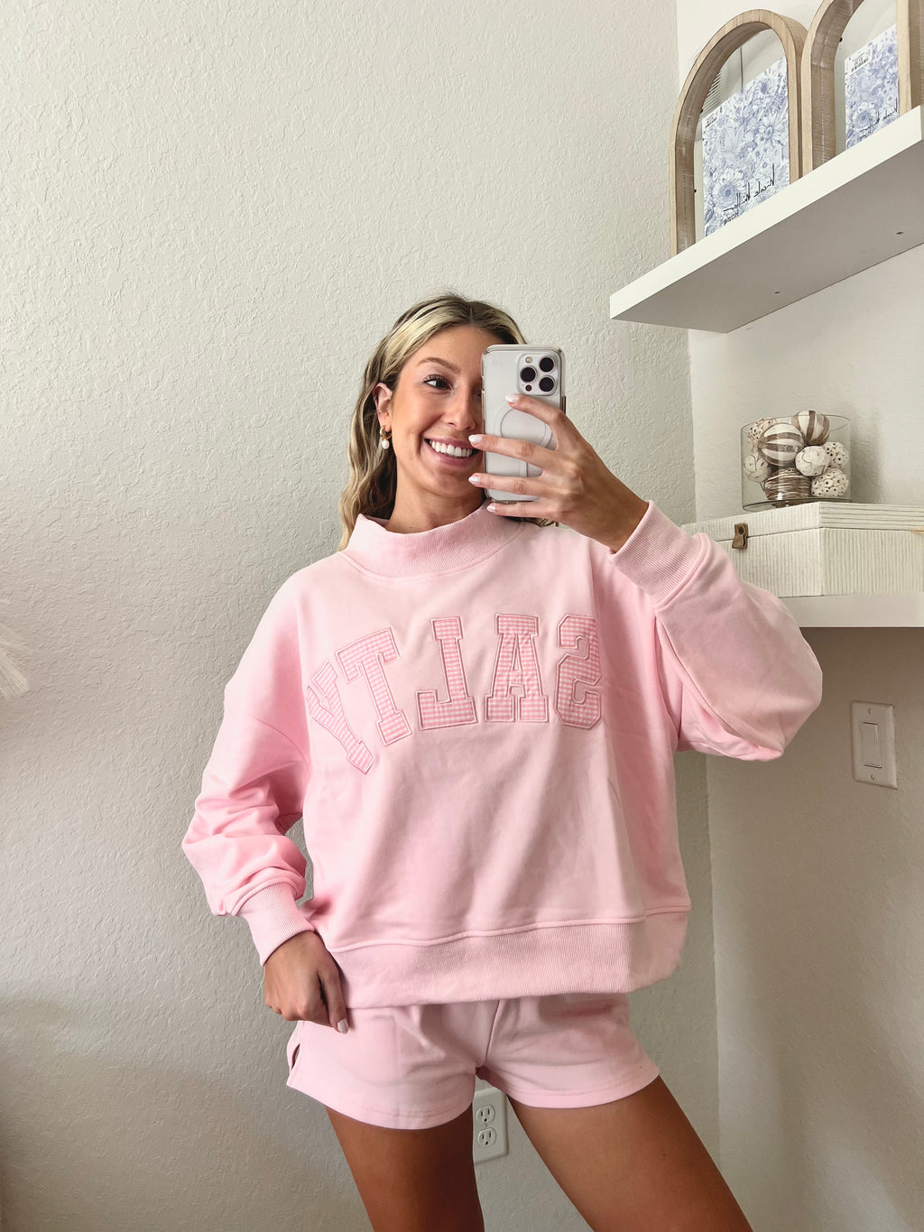 Woman wearing a pink sweatshirt with the word "Salty" on it and shorts set in a room with a shelf in the background.