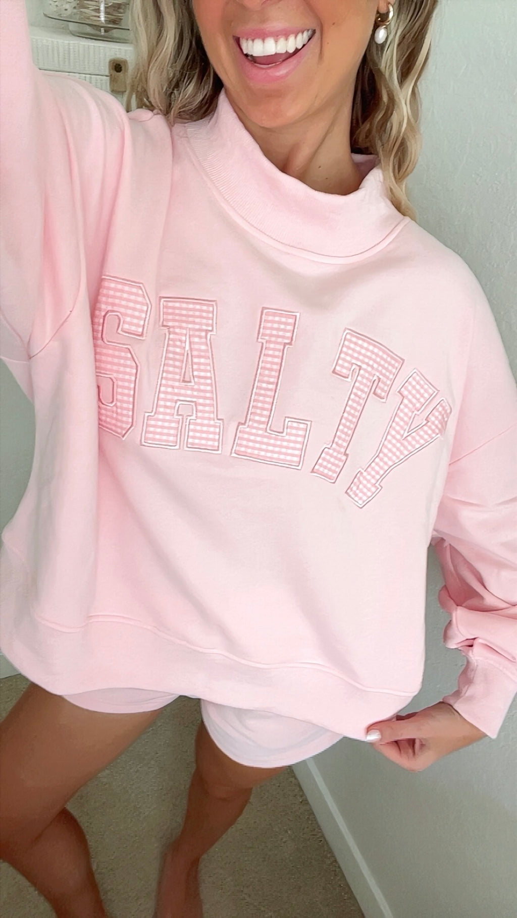 Woman wearing a pink sweatshirt with 'SALT' text, standing indoors.
