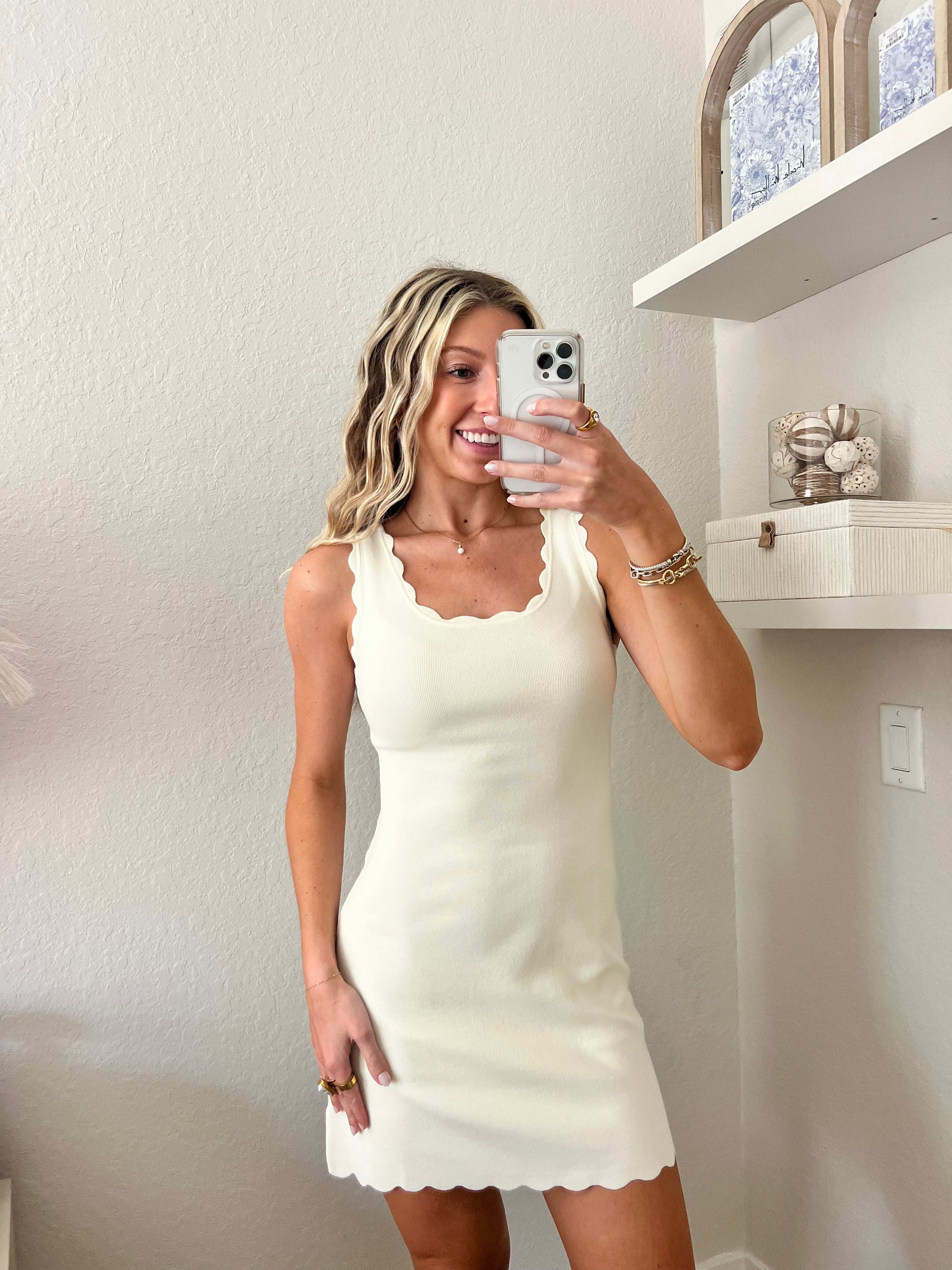 Woman taking a mirror selfie wearing an ivory dress in a room with a shelf and decorative items.