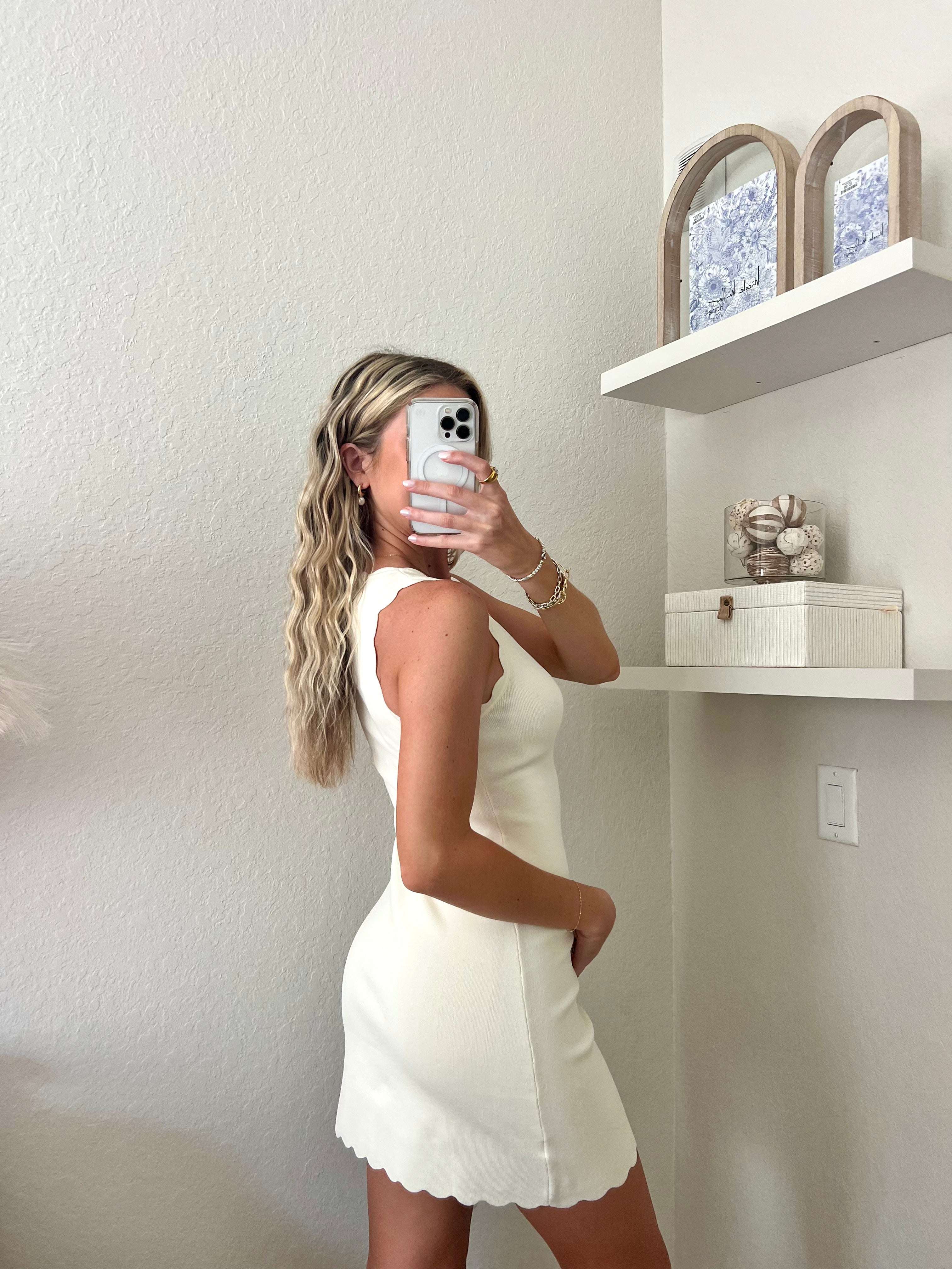 Woman taking a mirror selfie wearing an ivory dress in a room with a shelf and decorative items.