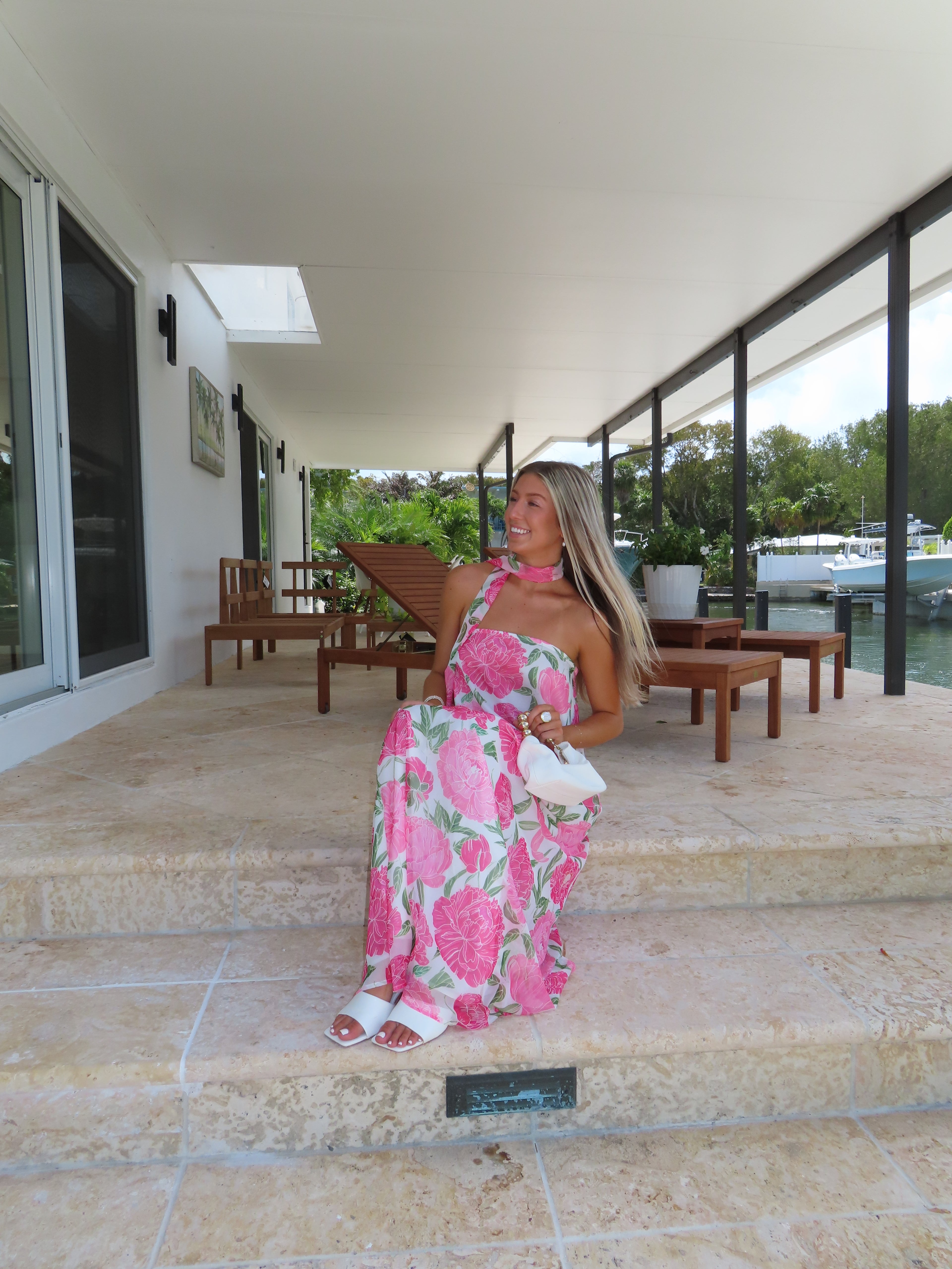 Woman in a pink floral maxi dress standing on a patio with tables and chairs.