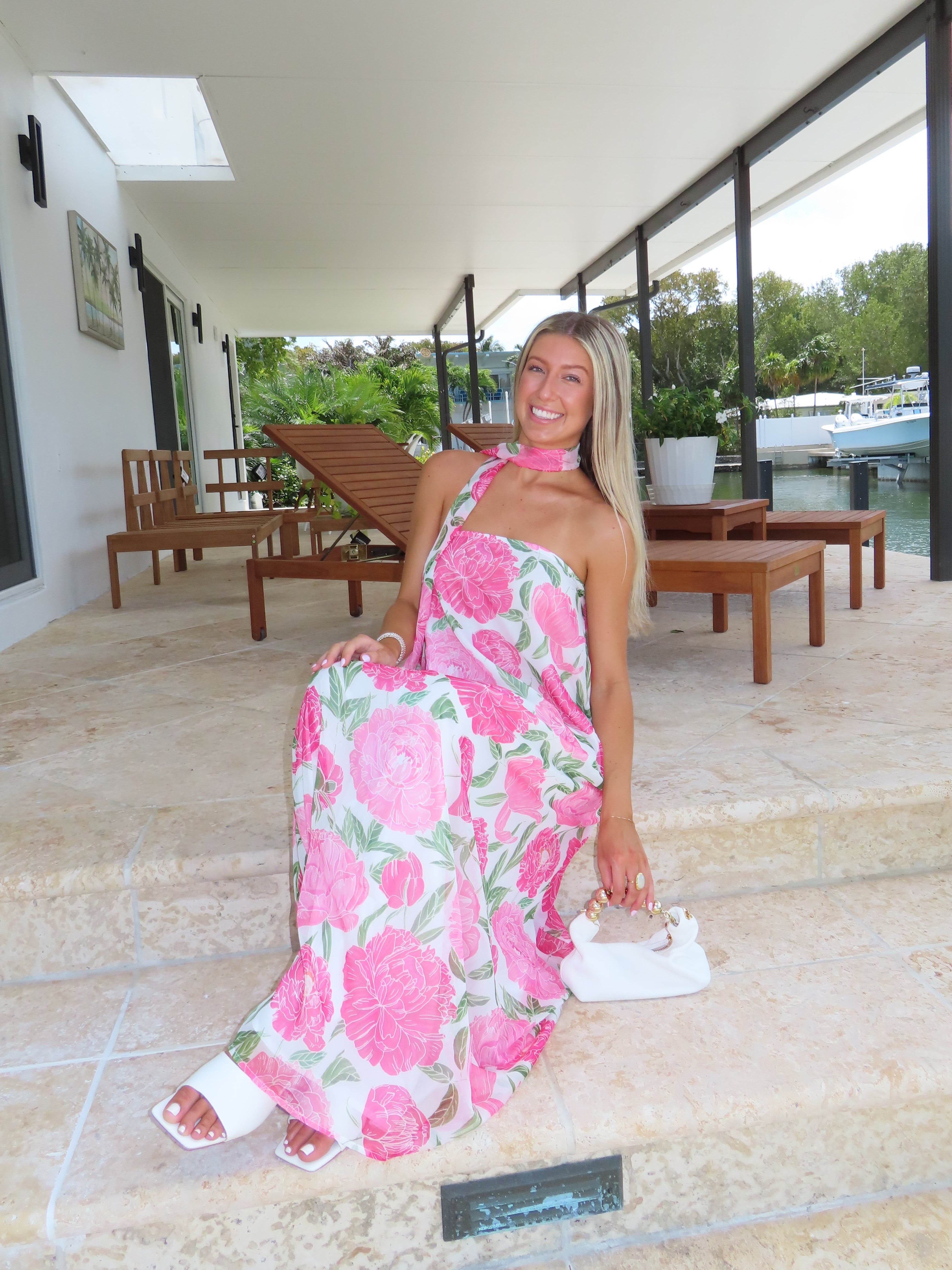 Woman in a pink floral maxi dress standing on a stone patio with greenery in the background
