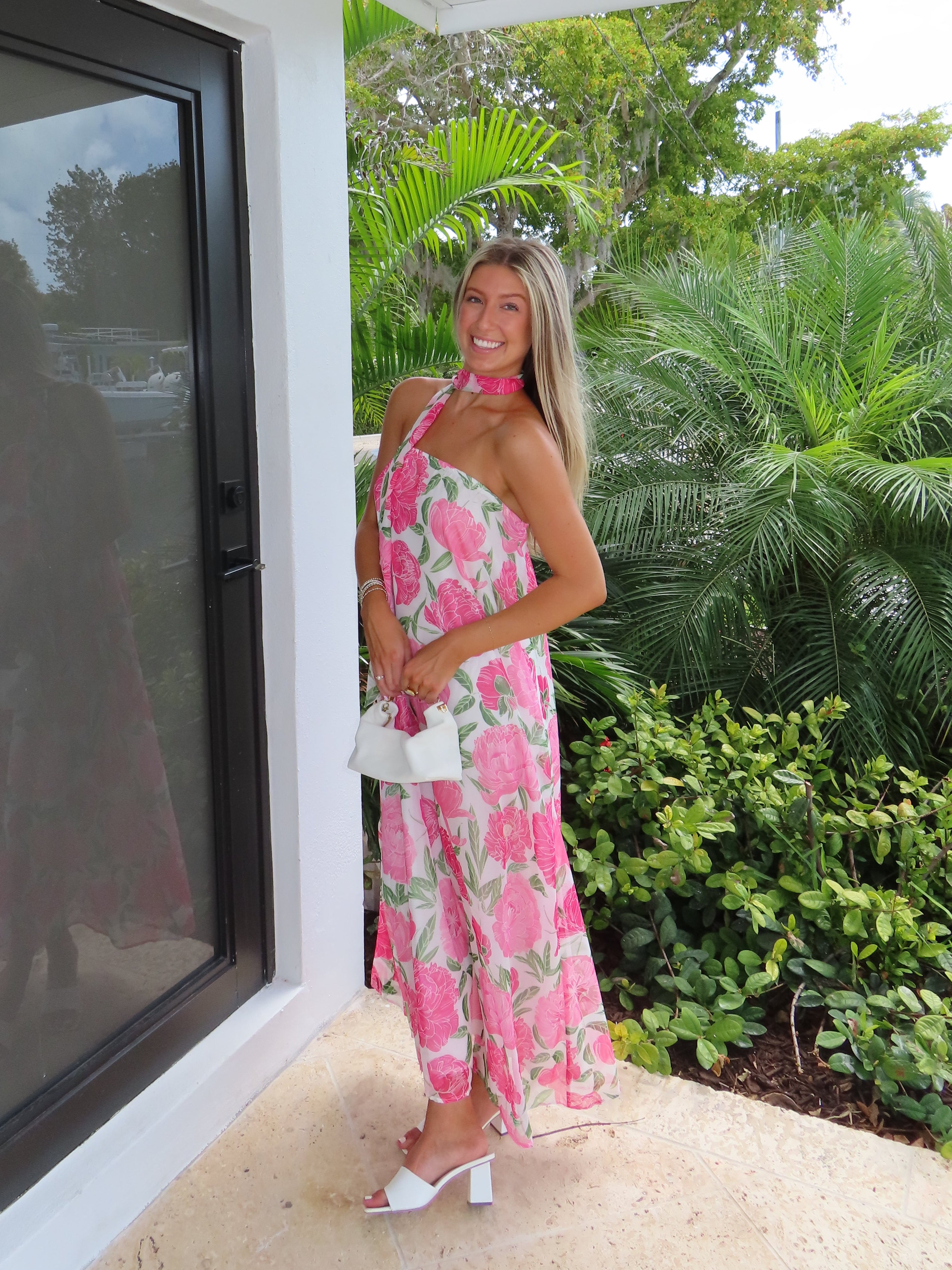 Woman in a pink floral maxi dress with detachable scarf standing next to a modern glass door with greenery outside.