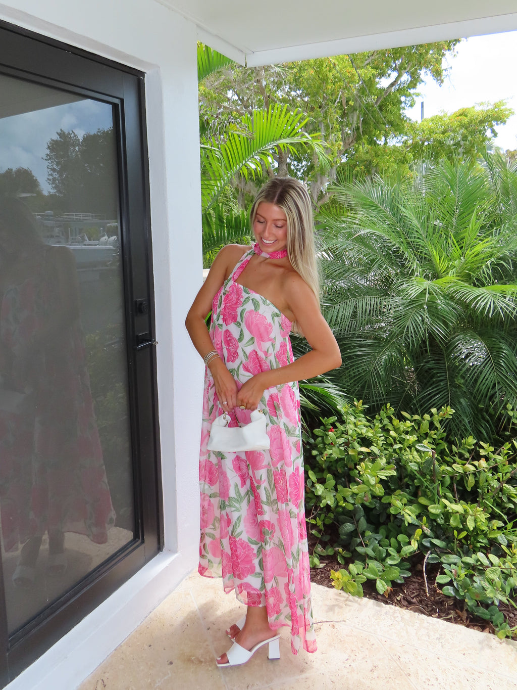Woman in a pink floral maxi dress with a scarf standing next to a modern outdoor structure with greenery in the background