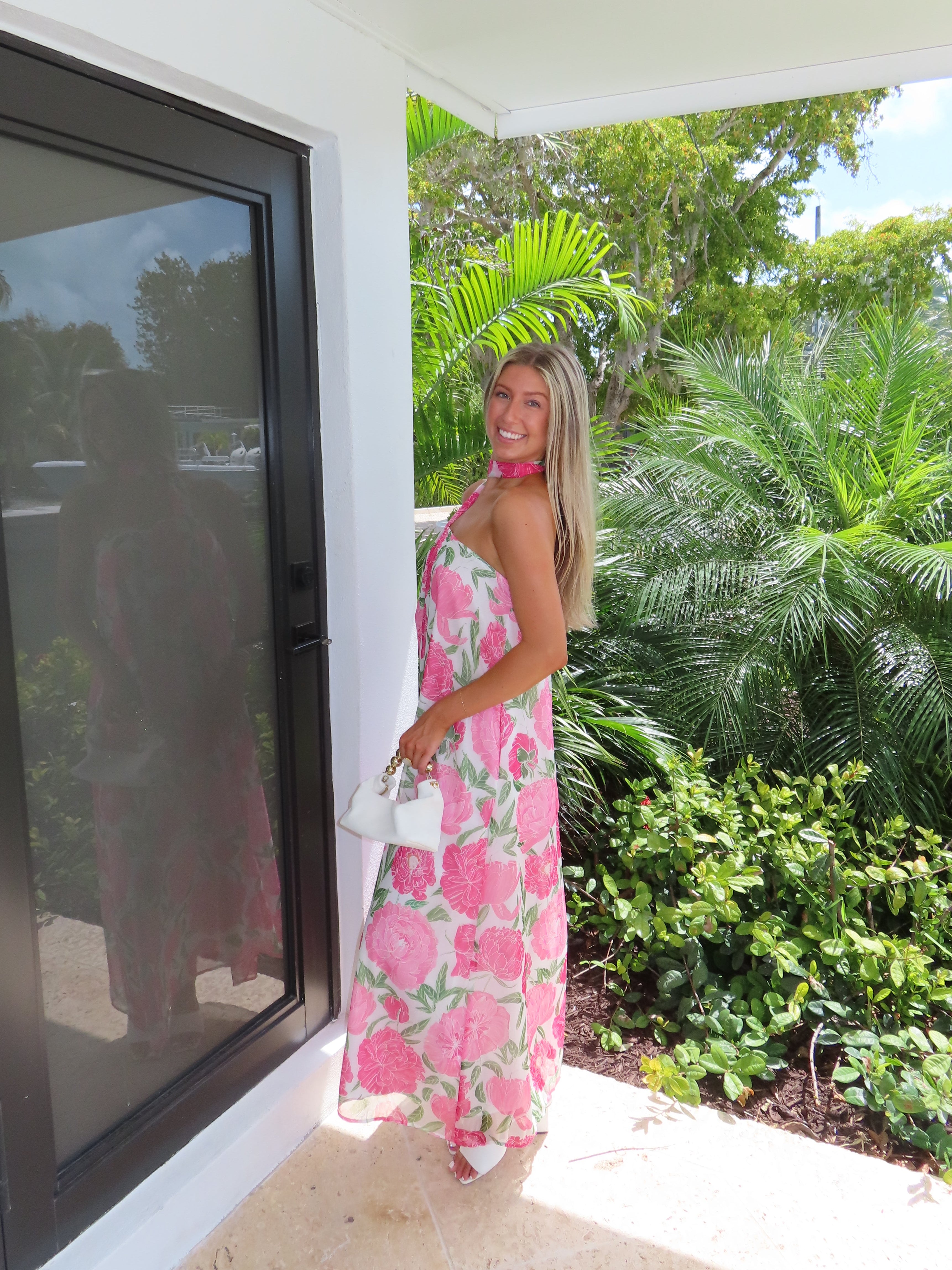 Woman in a pink floral maxi dress with a scarf standing next to a modern outdoor structure with greenery in the background