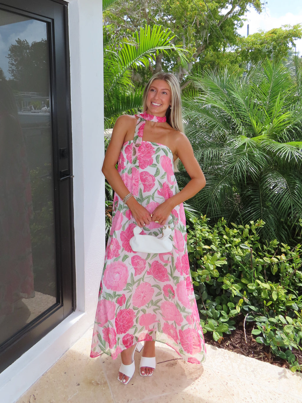 Woman in a pink floral maxi dress with a scarf standing next to a modern outdoor structure with greenery in the background