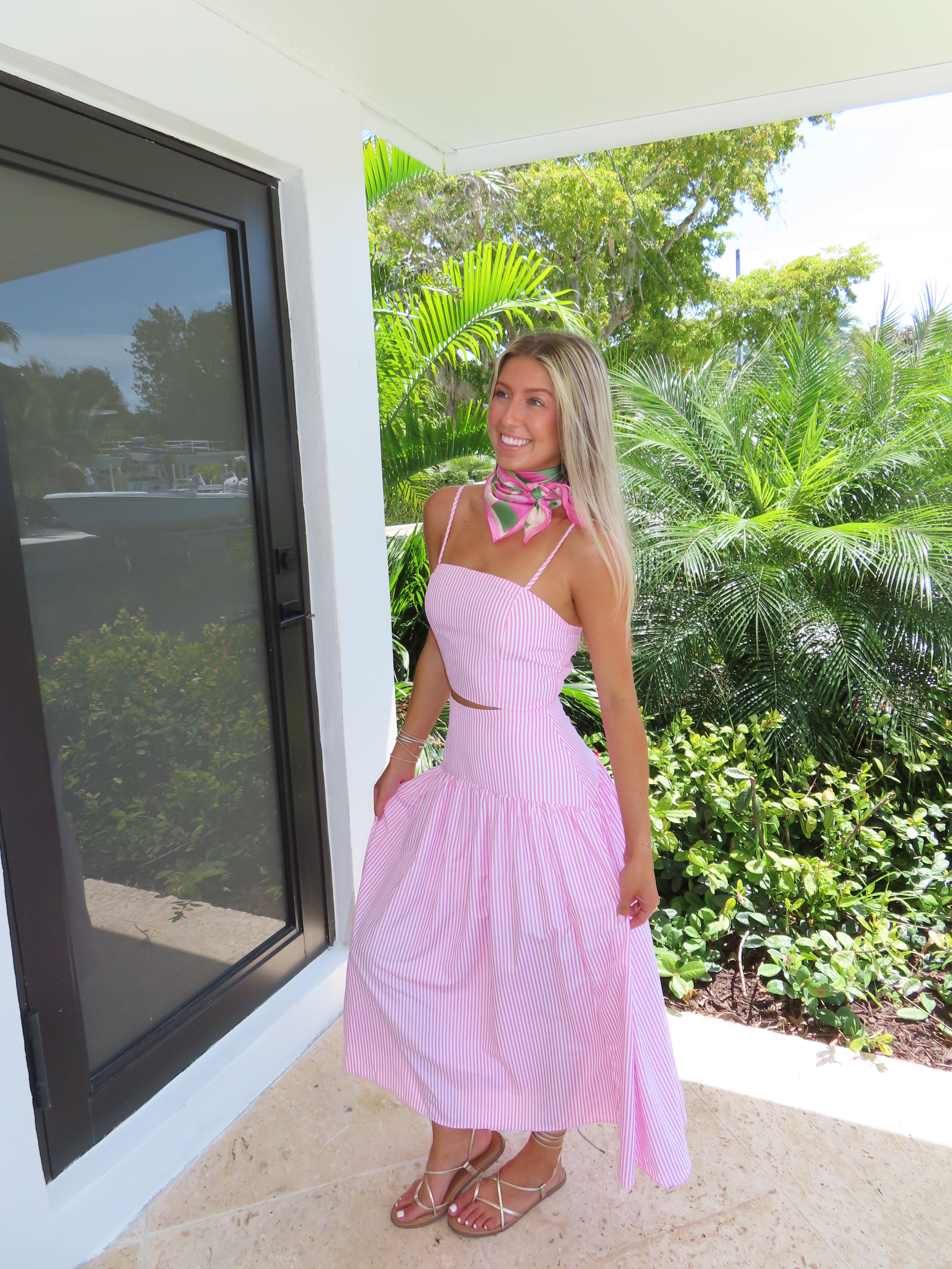 Woman in a pink and white striped maxi skirt set with a scarf standing outdoors with greenery in the background