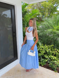 Woman in a white top and blue and white polka dot maxi skirt holding a white purse standing next to a glass door with greenery in the background