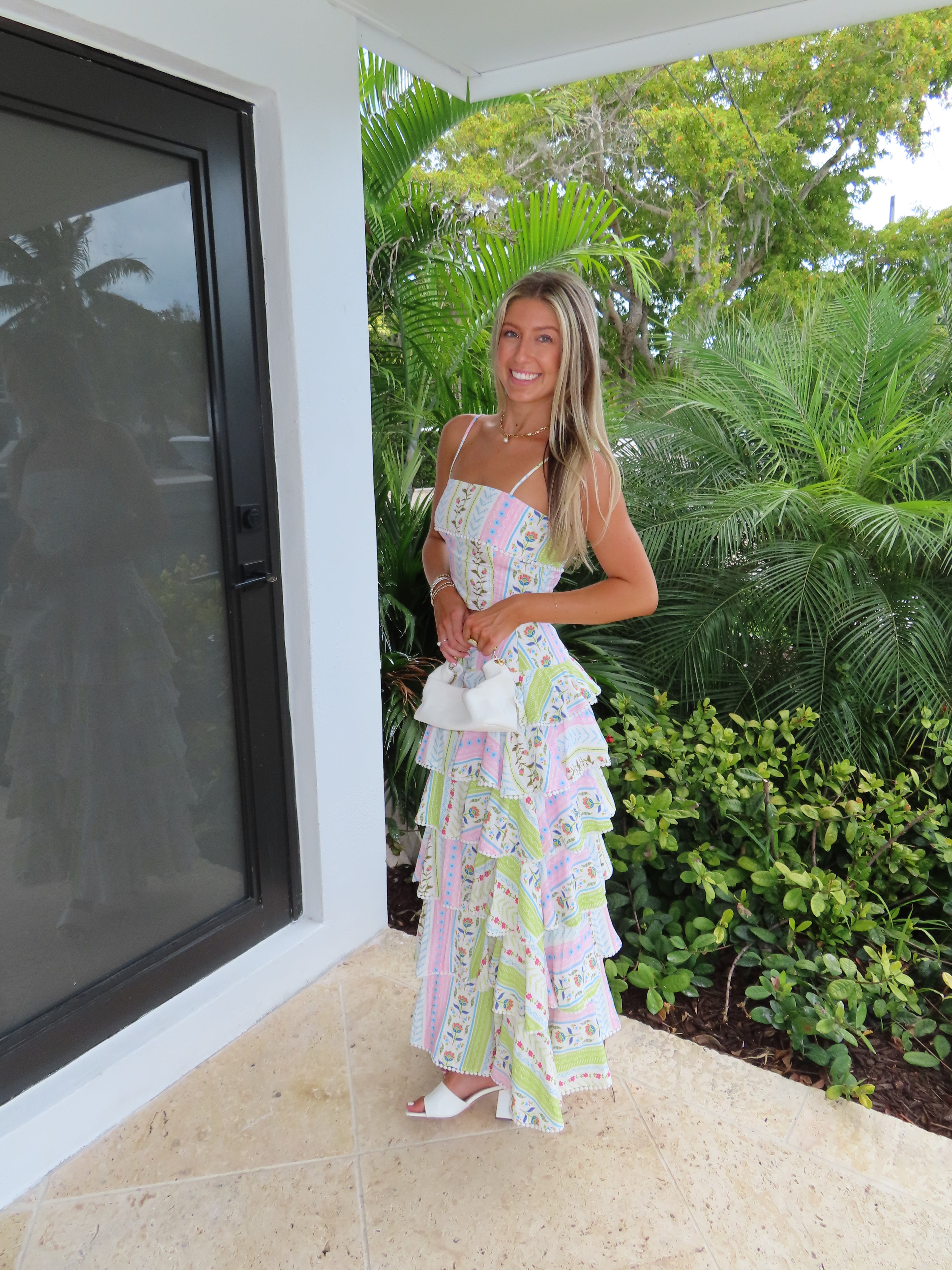 Woman holding a white purse in a patterned maxi skirt set standing near a glass door with greenery outside.